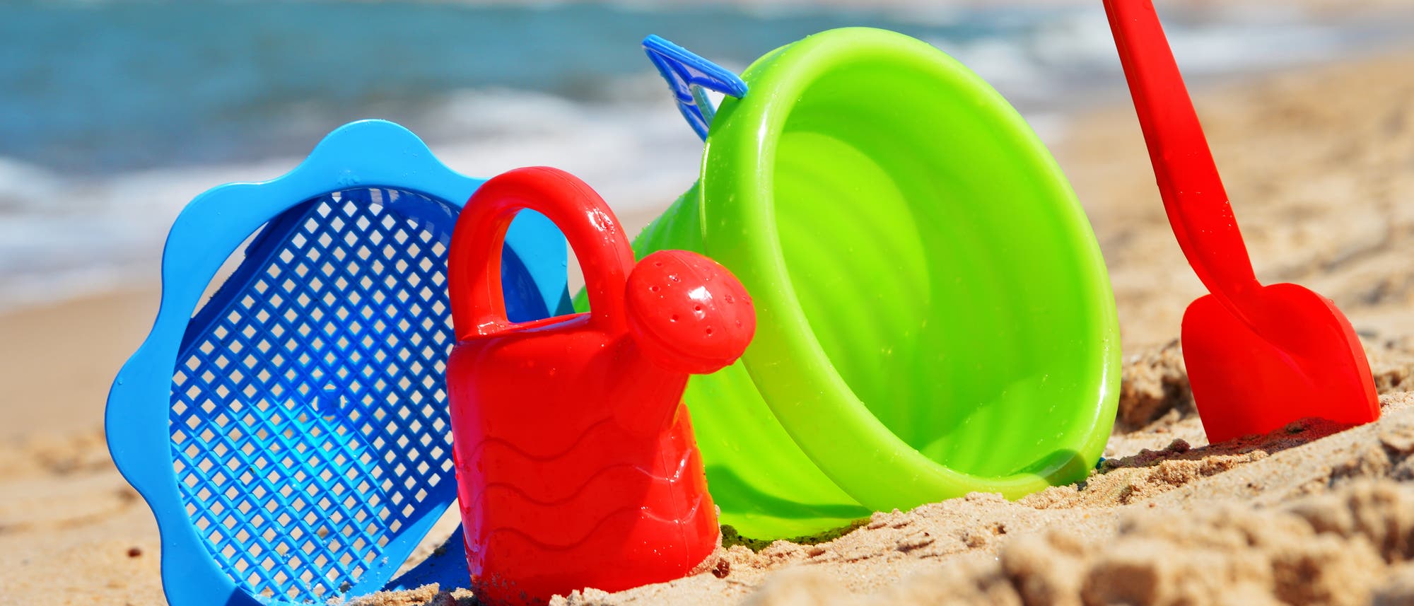 Sieb, Gießkanne, Eimer und Schaufel aus grellbuntem Plastik stehen im Sand an einem Strand