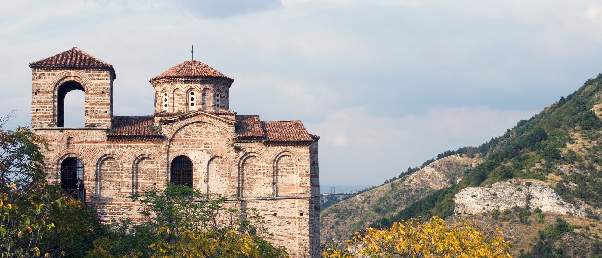 Eine alte, aus Ziegeln gebaute Kirche mit einem Turm und einer Kuppel steht auf einem bewaldeten H&uuml;gel. Im Hintergrund sind bewaldete Berge und ein bew&ouml;lkter Himmel zu sehen. Die Kirche ist von herbstlich gef&auml;rbten B&auml;umen umgeben.