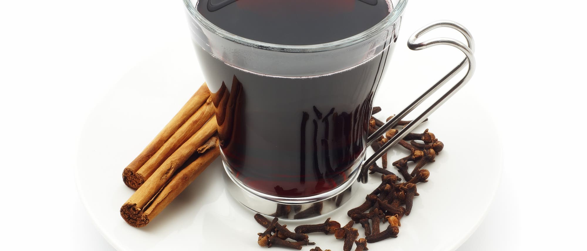 Ein Glas mit rotem Glühwein auf einem weißen Teller, umgeben von Zimtstangen und Nelken. Der Glühwein hat eine dunkle, satte Farbe und wird in einem Glas mit Metallgriff serviert. Die Gewürze auf dem Teller deuten auf die aromatische Zubereitung des Getränks hin. Ein Glas mit rotem Glühwein auf einem weißen Teller, umgeben von Zimtstangen und Nelken. Der Glühwein hat eine dunkle, satte Farbe und wird in einem Glas mit Metallgriff serviert. Die Gewürze auf dem Teller deuten auf die aromatische Zubereitung des Getränks hin.