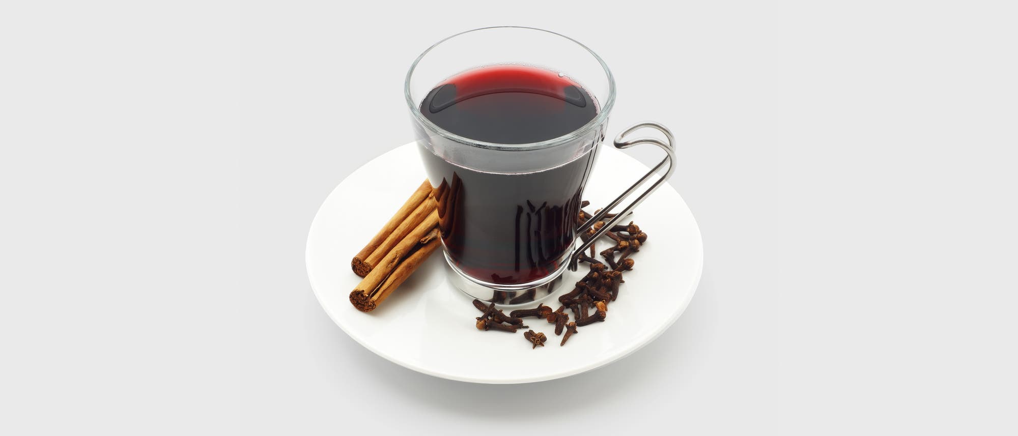 Ein Glas mit rotem Glühwein, umgeben von Zimtstangen und Nelken, auf einem grauen Hintergrund. Das Glas hat einen Metallgriff. 
