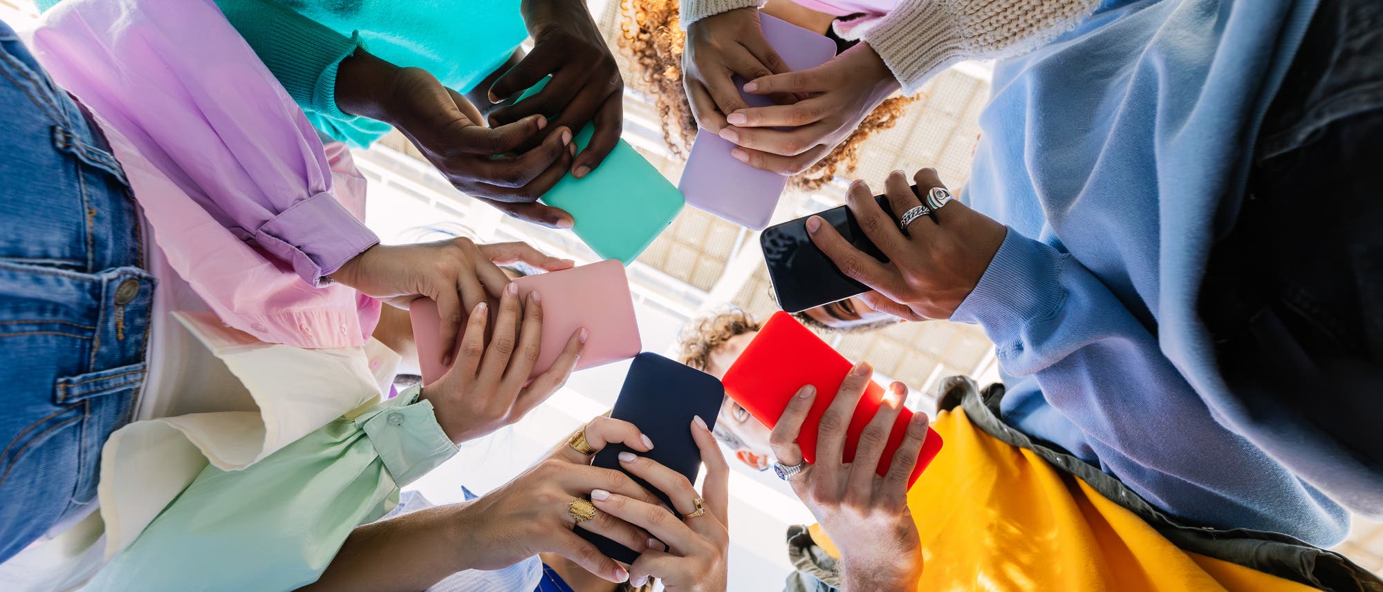 Eine Gruppe von Menschen steht im Kreis und h&auml;lt bunte Smartphones in den H&auml;nden. Die Aufnahme erfolgt von unten, sodass die Telefone im Mittelpunkt stehen. Die Personen tragen farbenfrohe Kleidung, und der Fokus liegt auf der gemeinsamen Nutzung von Technologie.