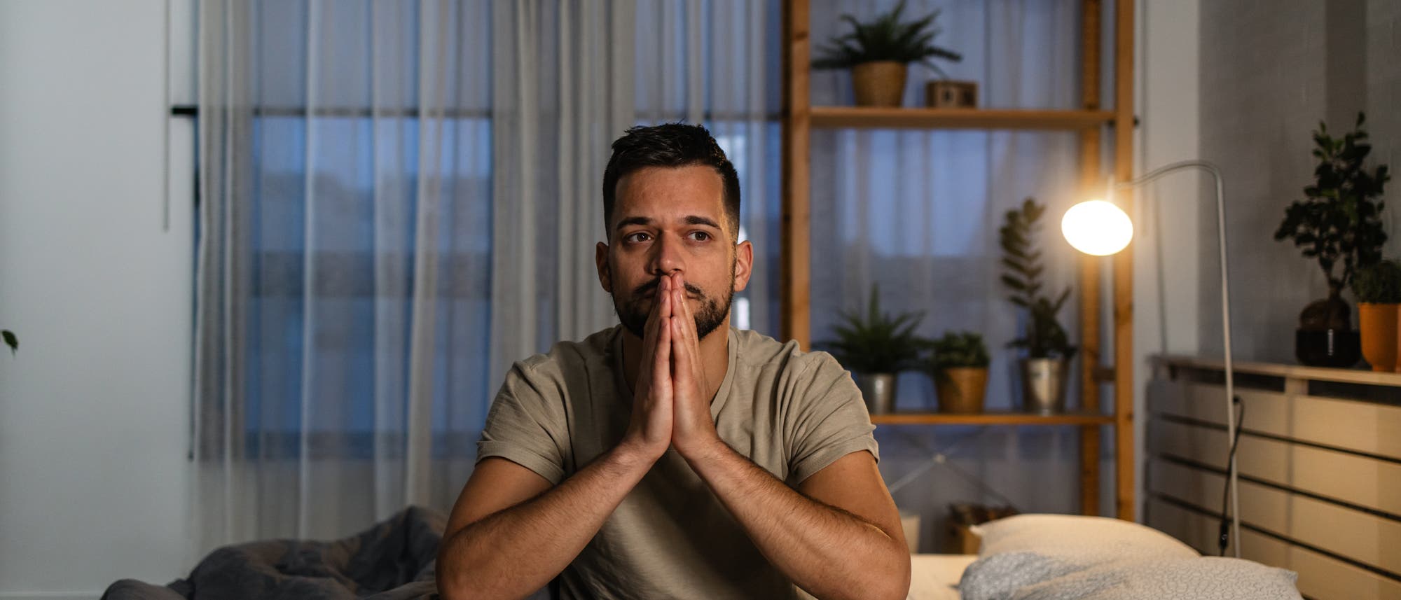 Ein Mann sitzt nachdenklich auf einem Bett in einem Schlafzimmer. Er trägt ein beiges T-Shirt und karierte Pyjamahosen. Im Hintergrund sind ein Regal mit Pflanzen und eine leuchtende Stehlampe zu sehen. Der Raum ist durch einen Vorhang abgedunkelt.