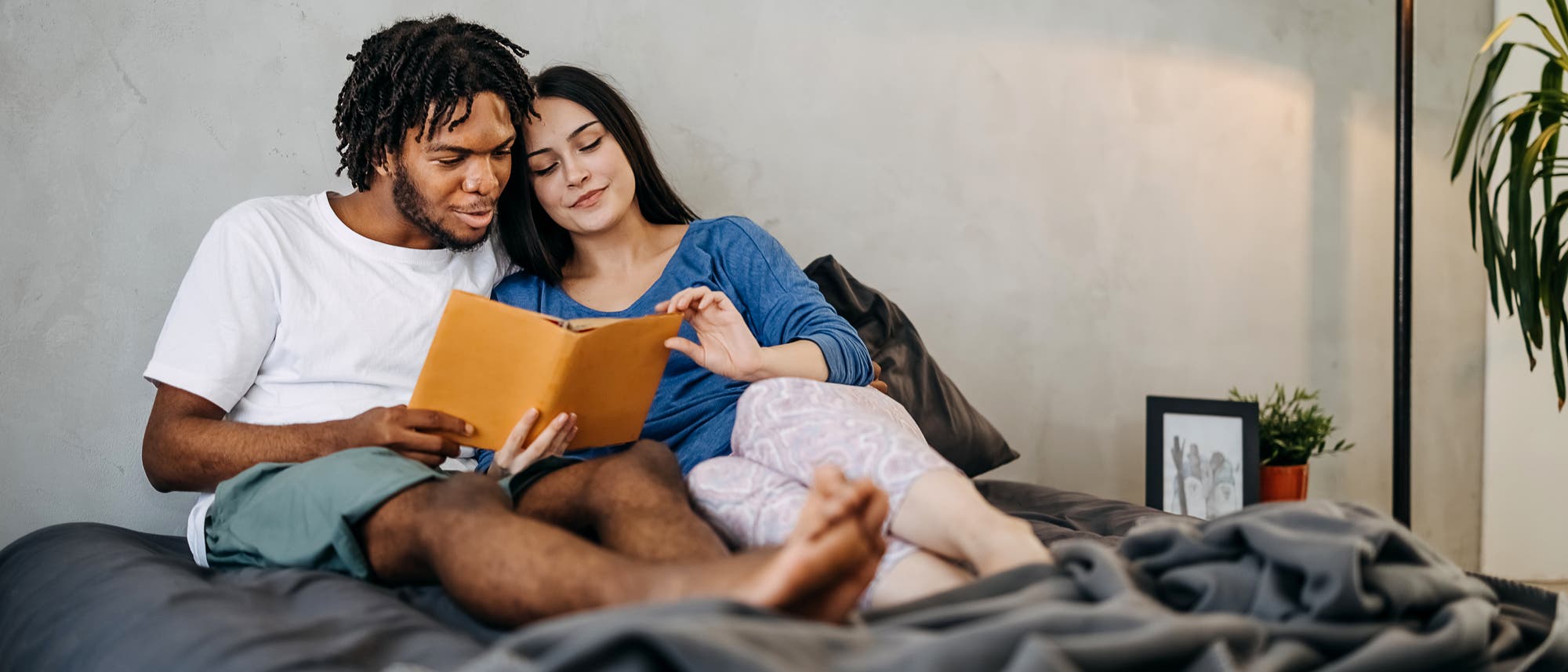 Ein Paar sitzt gemütlich auf einem Bett und liest gemeinsam ein Buch. Der Mann trägt ein weißes T-Shirt und Shorts, die Frau ein blaues Oberteil und gemusterte Pyjamahosen. Neben ihnen steht eine Stehlampe, auf dem Nachttisch stehen ein gerahmtes Foto sowie eine kleine Pflanze. Die Atmosphäre wirkt entspannt und harmonisch. Ein Paar sitzt gemütlich auf einem Bett und liest gemeinsam ein Buch. Der Mann trägt ein weißes T-Shirt und Shorts, die Frau ein blaues Oberteil und gemusterte Pyjamahosen. Neben ihnen steht eine Stehlampe, auf dem Nachttisch stehen ein gerahmtes Foto sowie eine kleine Pflanze. Die Atmosphäre wirkt entspannt und harmonisch.