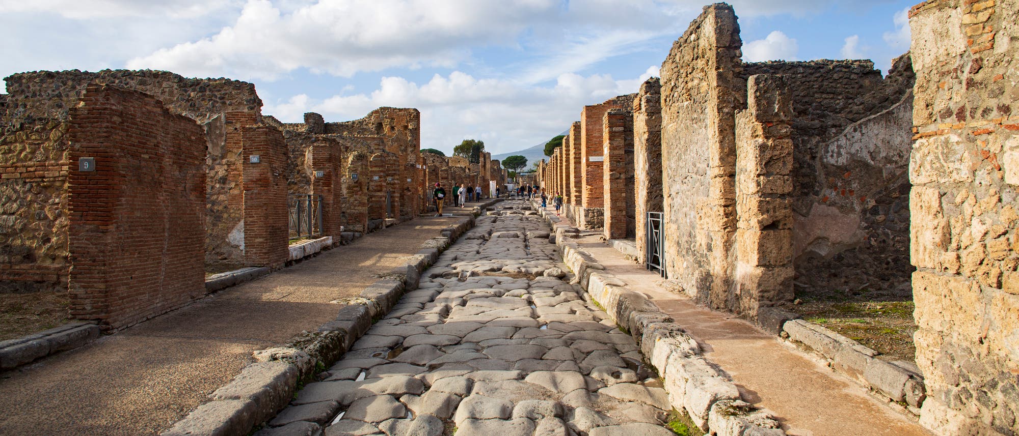 Eine antike, gepflasterte Straße in Pompeji, gesäumt von Ruinen aus Stein und Ziegeln. Der Himmel ist teils bewölkt, und einige Menschen sind in der Ferne zu sehen, die die historische Stätte erkunden. Die Szene vermittelt ein Gefühl von Geschichte und archäologischer Bedeutung. Eine antike, gepflasterte Straße in Pompeji, gesäumt von Ruinen aus Stein und Ziegeln. Der Himmel ist teils bewölkt, und einige Menschen sind in der Ferne zu sehen, die die historische Stätte erkunden. Die Szene vermittelt ein Gefühl von Geschichte und archäologischer Bedeutung.