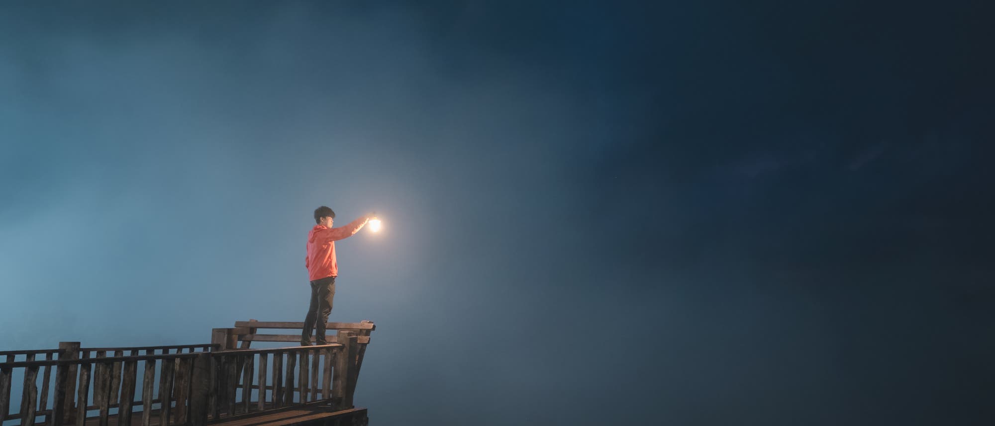 Eine Person steht auf einem Holzsteg und hält eine leuchtende Laterne in der Hand. Der Hintergrund ist dunkel und neblig, was eine mystische Atmosphäre erzeugt. Die Person trägt eine rote Jacke und blickt in die Ferne.