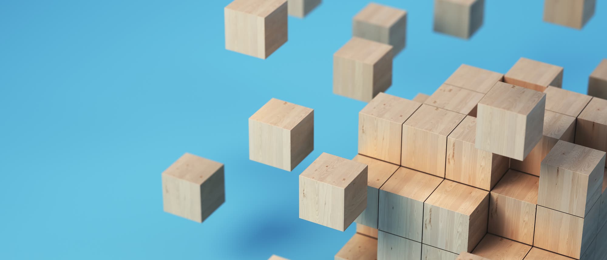 Eine 3D-Darstellung von hölzernen Würfeln, die in einem Raster angeordnet sind. Einige Würfel scheinen sich vom Hauptblock zu lösen und schweben in der Luft. Der Hintergrund ist einfarbig blau, was den Fokus auf die geometrische Anordnung der Würfel lenkt.