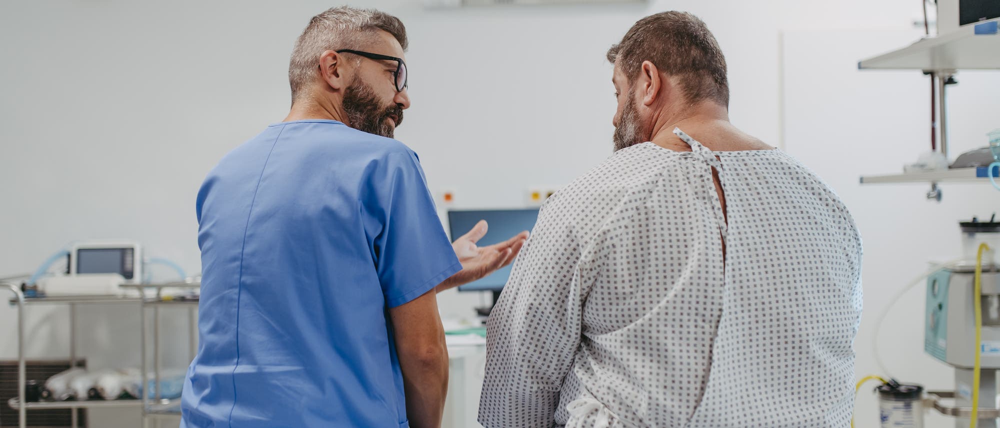 Ein Arzt in blauer Kleidung spricht mit einem Patienten mit Mehrgewicht in einem Krankenhauskittel. Beide sitzen auf einem Untersuchungstisch in einem medizinischen Behandlungsraum. Im Hintergrund sind medizinische Ger&auml;te und eine rote Uhr zu sehen. Der Arzt gestikuliert, w&auml;hrend er mit dem Patienten spricht, was auf eine Beratung oder Erkl&auml;rung hindeutet.