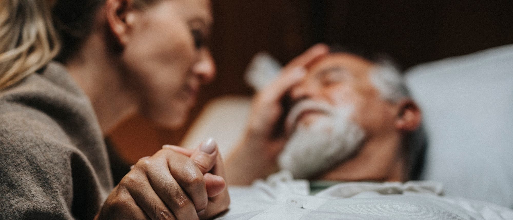Eine Frau h&auml;lt die Hand eines &auml;lteren Mannes, der im Bett liegt. Sie beugt sich zu ihm hin&uuml;ber und ber&uuml;hrt sanft seine Stirn. Die Szene vermittelt F&uuml;rsorge und Trost in einer ruhigen Umgebung.