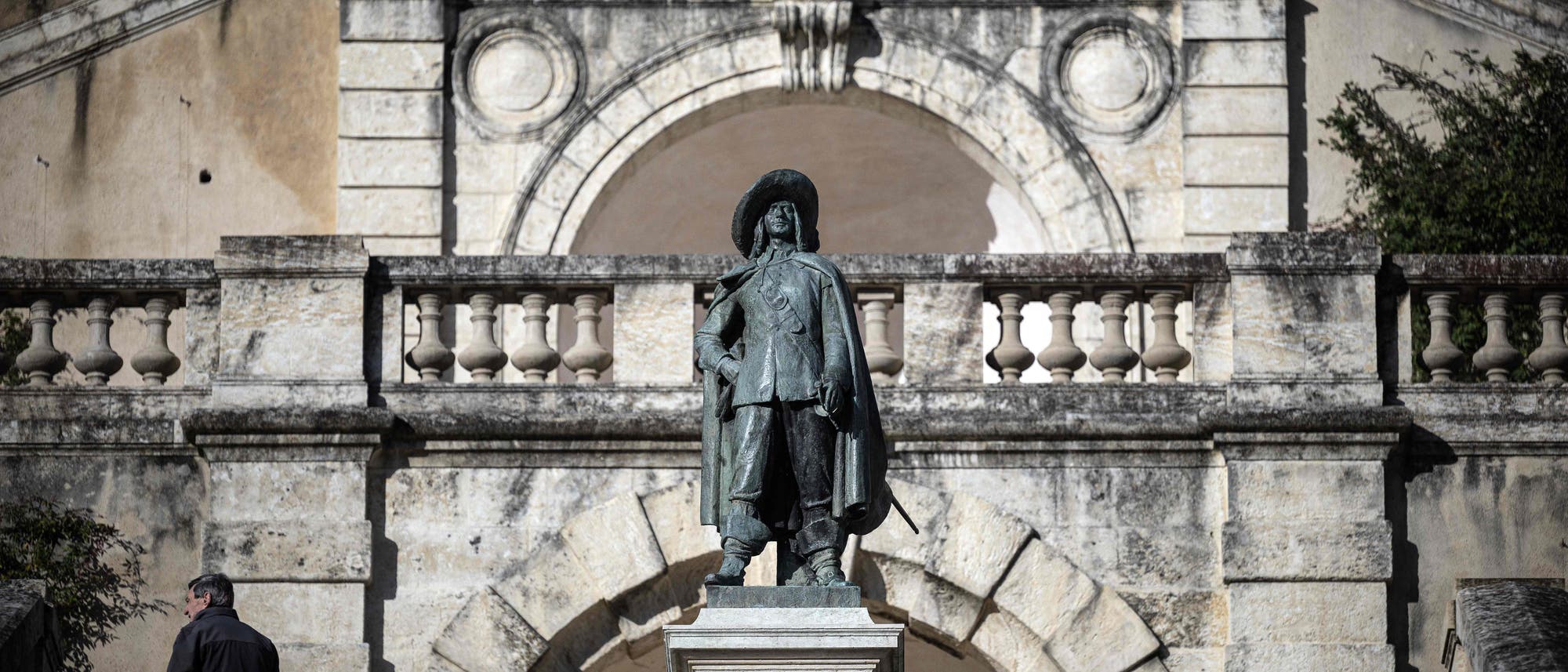 Eine Statue von D&rsquo;Artagnan steht vor einer historischen Steinarchitektur mit Balustraden und dekorativen Elementen. Eine Person geht links im Bild die Treppe hinauf. Die Szene ist bei Tageslicht aufgenommen.