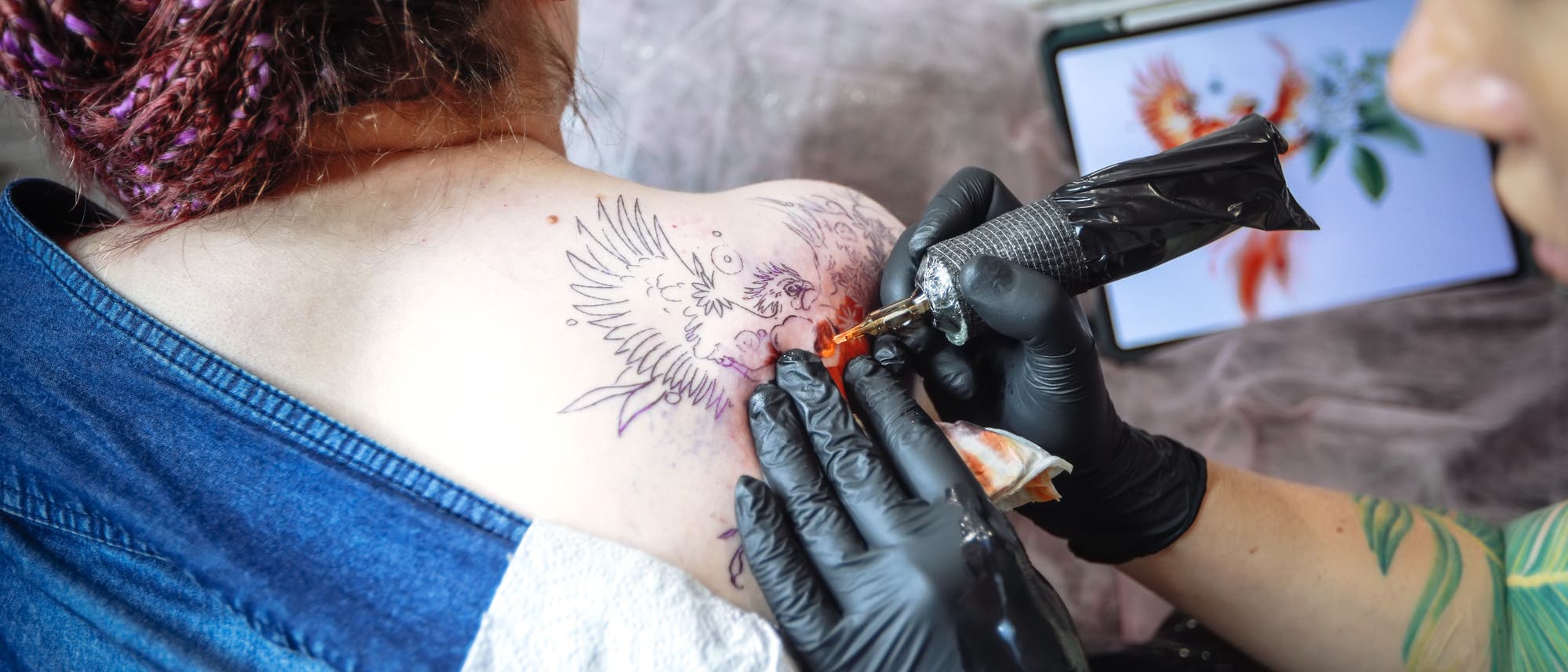 Eine Person erh&auml;lt ein Tattoo auf der Schulter, das einen Vogel mit ausgebreiteten Fl&uuml;geln zeigt. Der T&auml;towierer tr&auml;gt schwarze Handschuhe und verwendet eine T&auml;towiermaschine. Im Hintergrund ist ein Tablet mit einer farbigen Darstellung des Vogels zu sehen. Die Szene findet in einem Studio mit einer wei&szlig;en Backsteinwand statt.