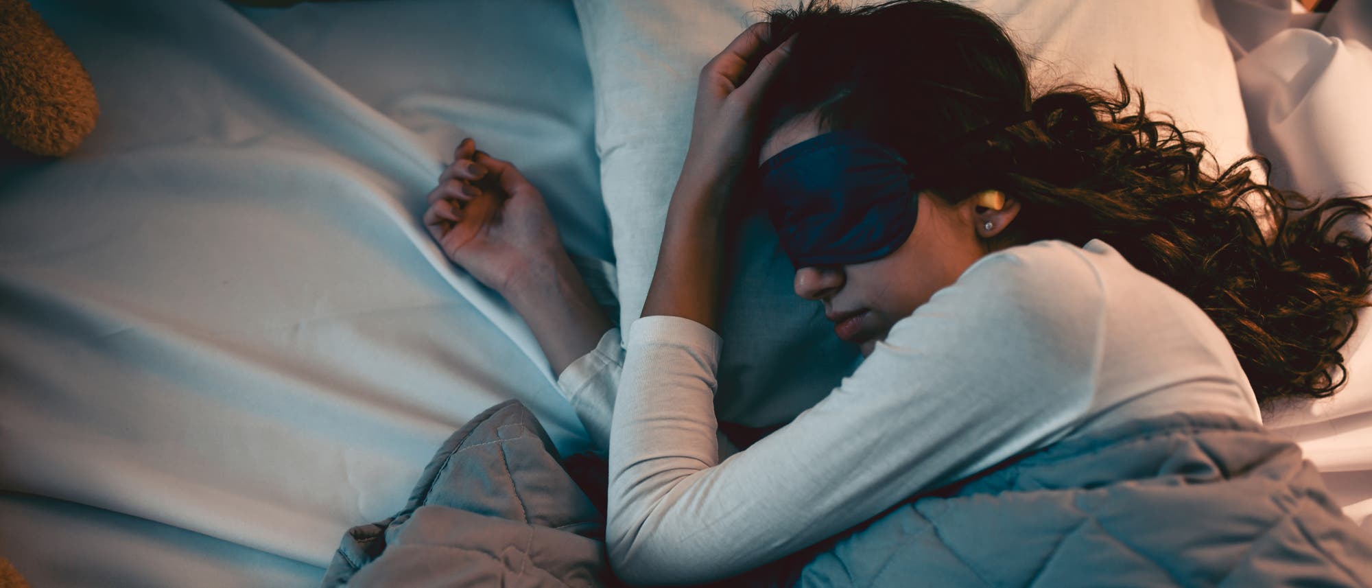 Eine Person liegt schlafend in einem Bett, zugedeckt mit einer grauen Decke. Sie tr&auml;gt eine Schlafmaske und Ohrst&ouml;psel. Die Szene vermittelt Ruhe und Entspannung. Im Hintergrund sind ein Kissen und ein Pl&uuml;schtier zu sehen.