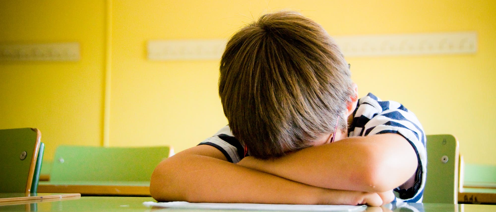 Ein Kind sitzt in einem Klassenzimmer an einem Tisch und hat den Kopf auf die verschr&auml;nkten Arme gelegt. Die Umgebung ist hell mit gelben W&auml;nden und gr&uuml;nen St&uuml;hlen. Das Bild vermittelt den Eindruck von M&uuml;digkeit, Langeweile oder Frustration.