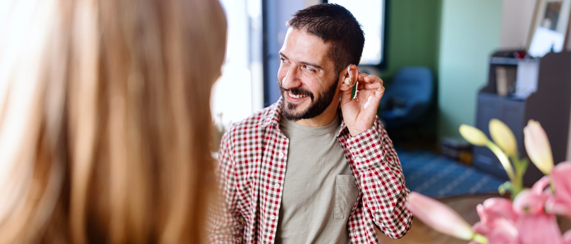 Ein Mann in einem karierten Hemd l&auml;chelt und zeigt auf sein H&ouml;rger&auml;t, w&auml;hrend er mit einer anderen Person spricht. Die Szene spielt in einem hellen Raum mit einem Fenster im Hintergrund. Das Bild vermittelt eine positive Interaktion und den Einsatz von H&ouml;rhilfen.