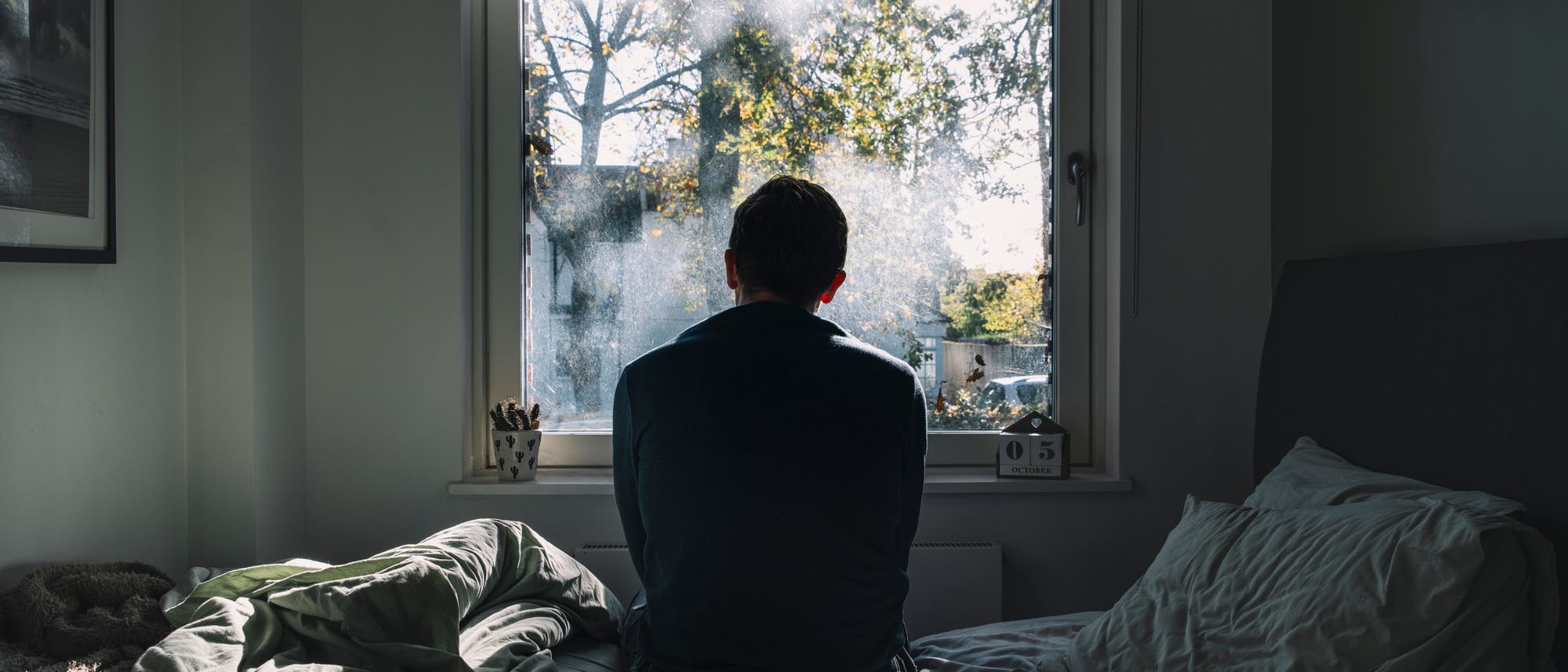 Eine Person sitzt auf einem Bett und blickt aus einem Fenster, durch das Sonnenlicht auf die Szene f&auml;llt. Drau&szlig;en sind B&auml;ume mit Herbstlaub zu sehen. Auf dem Fensterbrett steht ein Kalender, der den 5. Oktober anzeigt. Die Bettdecke ist unordentlich, und der Raum wirkt ruhig und nachdenklich.