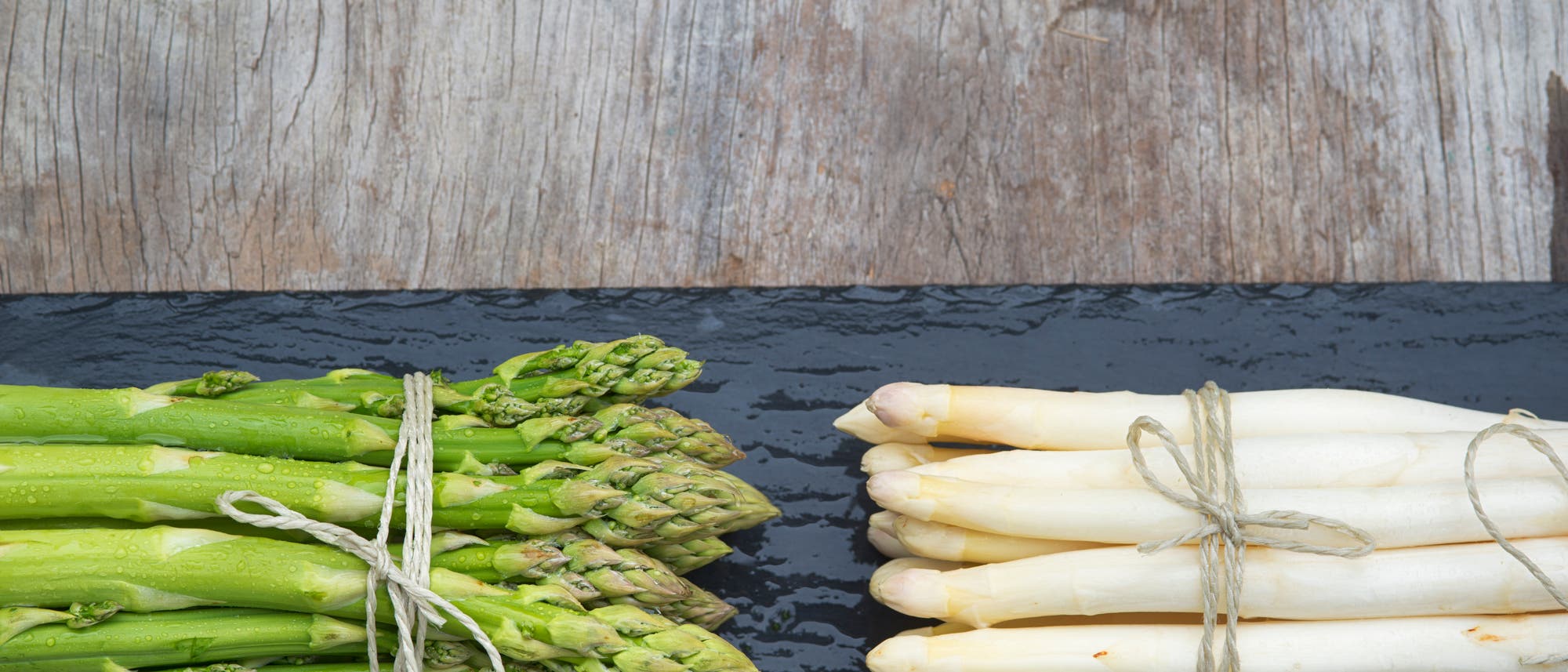 Ein B&uuml;ndel gr&uuml;ner Spargel und ein B&uuml;ndel wei&szlig;er Spargel liegen nebeneinander auf einer dunklen Schieferplatte. Beide B&uuml;ndel sind mit Schnur zusammengebunden. Der Hintergrund besteht aus einer rustikalen Holzoberfl&auml;che.