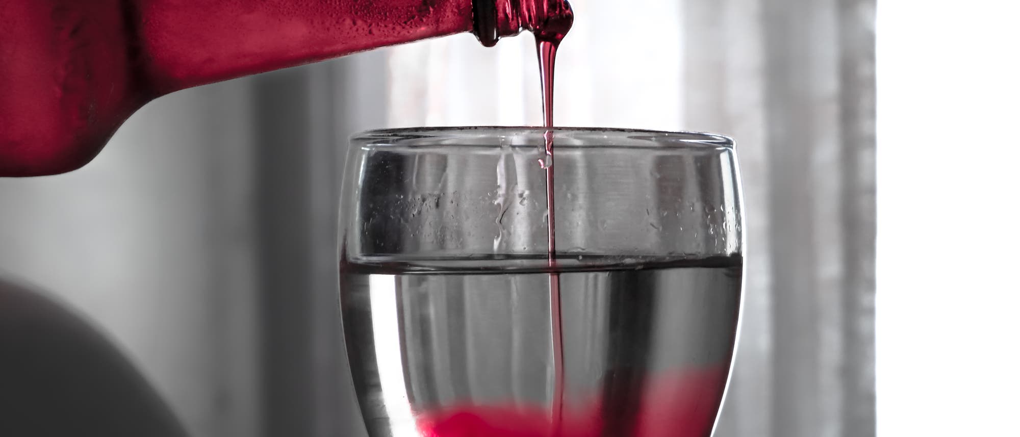 Eine Flasche mit rotem Getr&auml;nk wird in ein klares Glas gegossen. Der rote Fl&uuml;ssigkeitsstrom trifft auf das Wasser im Glas und erzeugt einen Farbverlauf. Im Hintergrund sind verschwommene graue Vorh&auml;nge zu sehen.