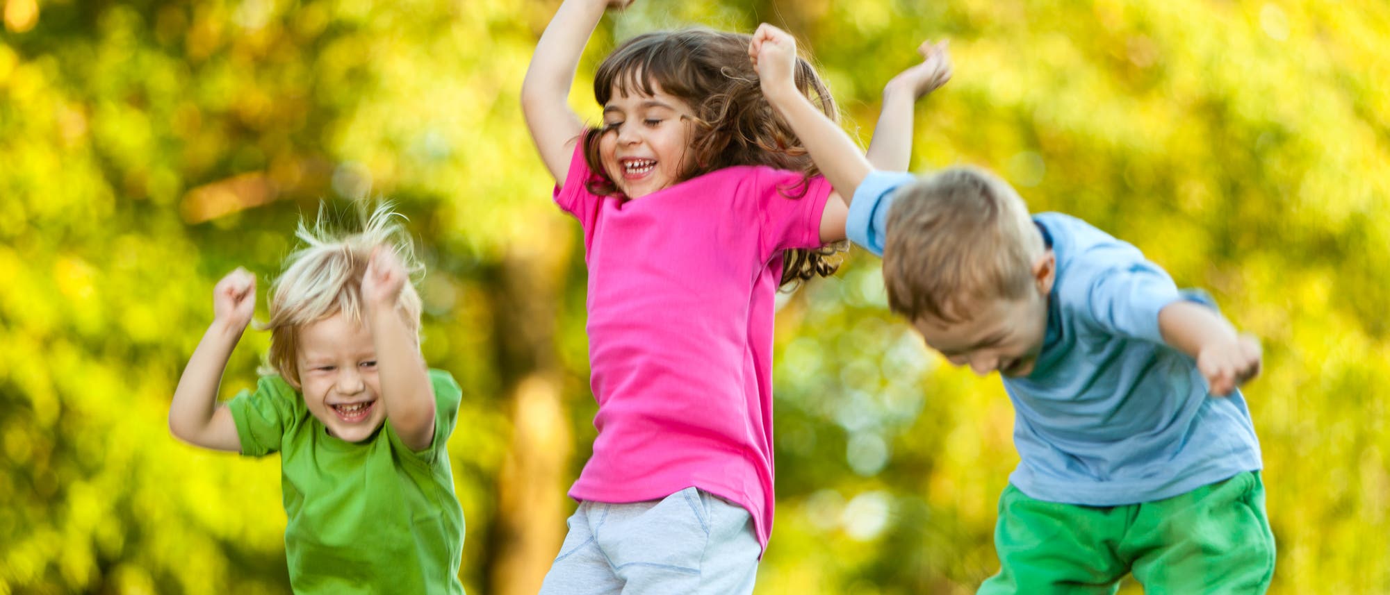 Drei kleine Kinder springen lachend unter freiem Himmel umher.