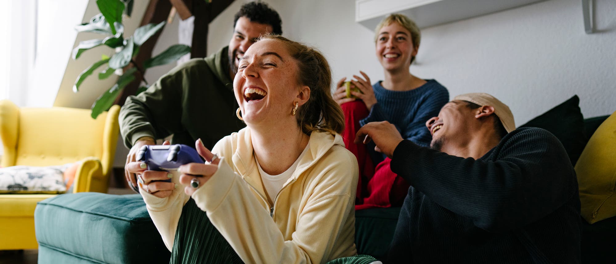 Eine Gruppe von vier Personen sitzt in einem Wohnzimmer auf einem Sofa und lacht, während sie Videospiele spielen. Zwei Personen halten Controller in den Händen. Die Atmosphäre ist entspannt und fröhlich. Im Hintergrund sind Pflanzen und ein gelber Sessel zu sehen. Eine Gruppe von vier Personen sitzt in einem Wohnzimmer auf einem Sofa und lacht, während sie Videospiele spielen. Zwei Personen halten Controller in den Händen. Die Atmosphäre ist entspannt und fröhlich. Im Hintergrund sind Pflanzen und ein gelber Sessel zu sehen.