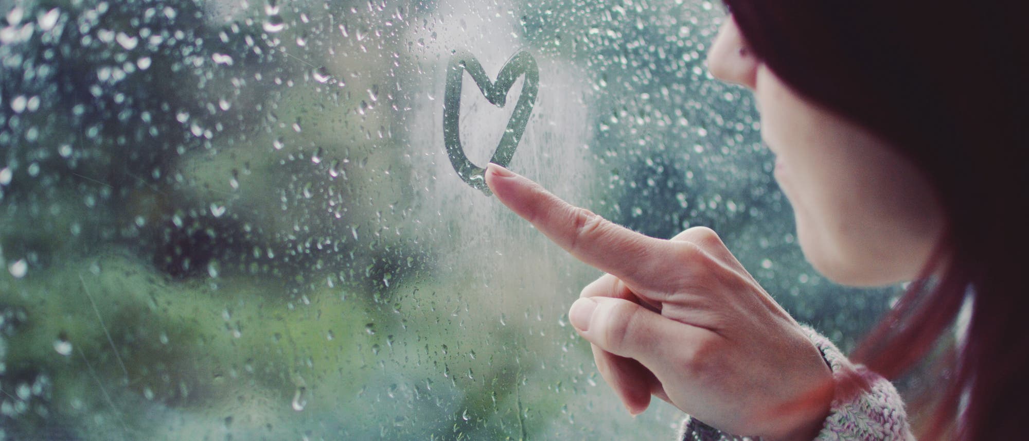 Eine Person zeichnet mit dem Finger ein Herz auf eine regennasse Fensterscheibe. Drau&szlig;en ist eine verschwommene, gr&uuml;ne Landschaft zu sehen. Die Szene vermittelt eine ruhige und nachdenkliche Stimmung.