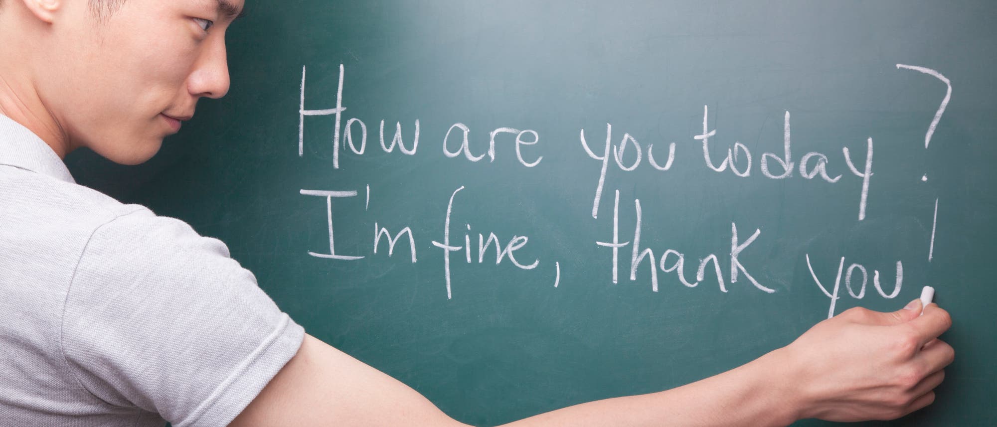 Ein Mann schreibt mit Kreide auf eine grüne Tafel. Der Text auf der Tafel lautet: "How are you today? I'm fine, thank you!" Der Mann trägt ein helles T-Shirt und konzentriert sich auf das Schreiben.