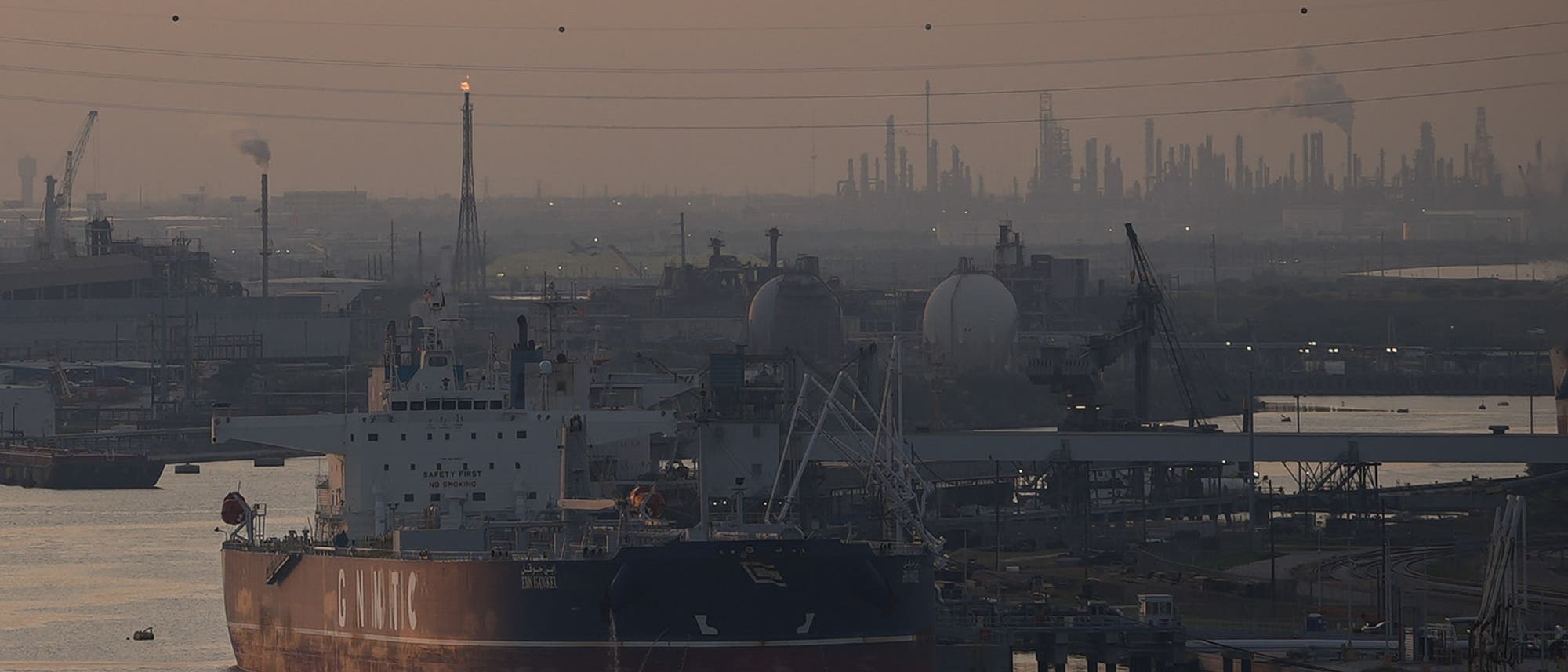 Ein großes Frachtschiff liegt in einem Industriehafen vor Anker. Im Hintergrund sind zahlreiche Fabrikanlagen und Schornsteine zu sehen, aus denen Rauch aufsteigt. Die Szene ist in ein gedämpftes Licht getaucht, das eine dunstige Atmosphäre erzeugt. Stromleitungen verlaufen über den Hafen, und Kräne sind entlang der Uferlinie sichtbar.
