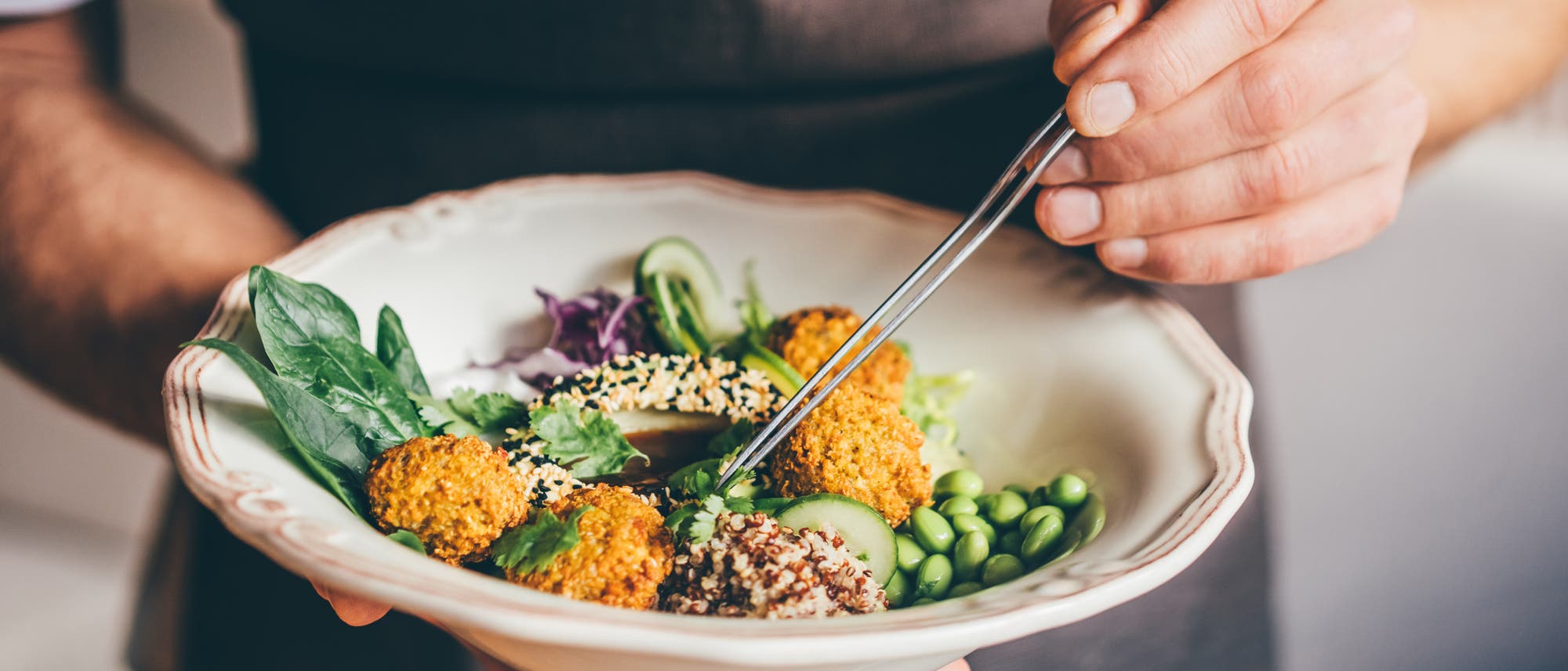 Eine Person in einer Schürze hält eine Schüssel mit einem bunten Salat, der aus Falafel, Quinoa, Edamame, Gurkenscheiben, Spinat und Sesam besteht. Die Person arrangiert die Zutaten mit einer Pinzette. Der Hintergrund ist unscharf, was den Fokus auf das Essen lenkt. Eine Person in einer Schürze hält eine Schüssel mit einem bunten Salat, der aus Falafel, Quinoa, Edamame, Gurkenscheiben, Spinat und Sesam besteht. Die Person arrangiert die Zutaten mit einer Pinzette. Der Hintergrund ist unscharf, was den Fokus auf das Essen lenkt.