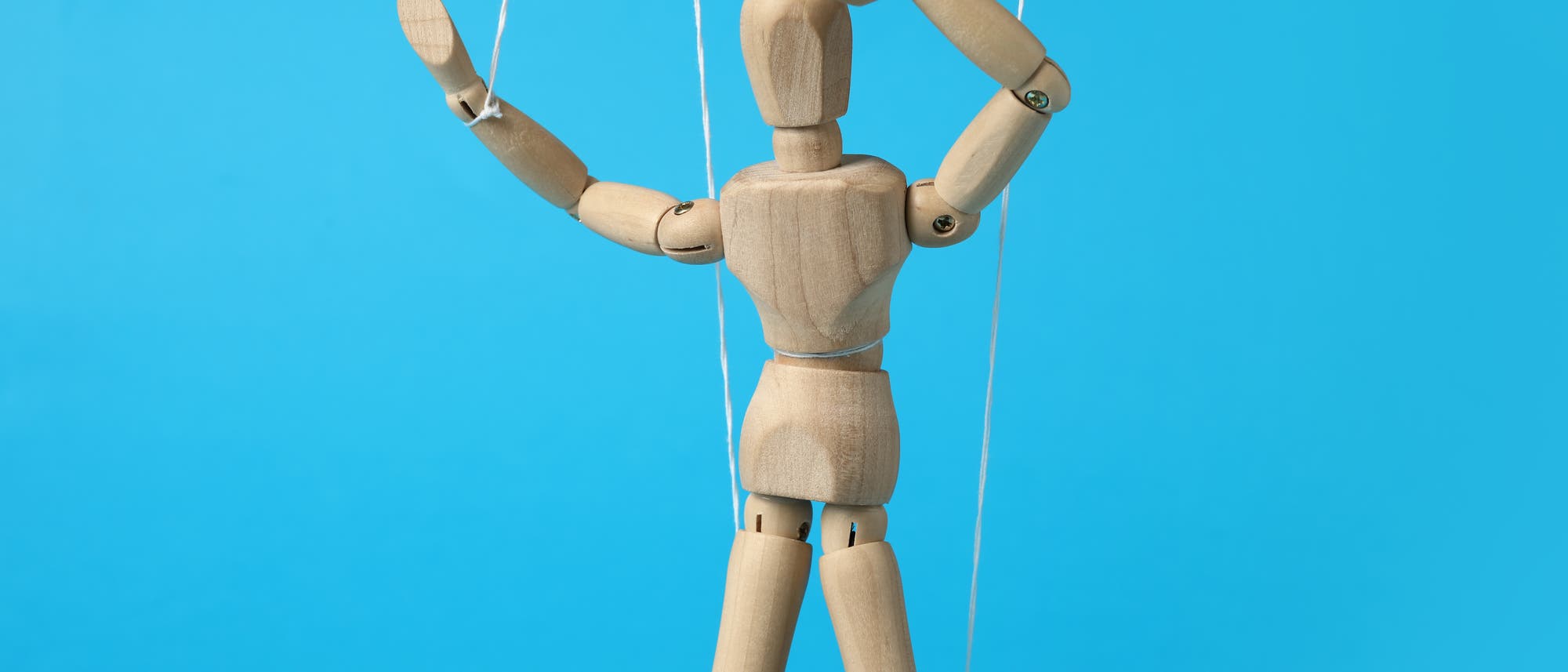 Eine hölzerne Gliederpuppe, die an Fäden aufgehängt ist, vor einem blauen Hintergrund. Die Puppe ist in einer Pose, die an eine Marionette erinnert, mit einem Arm erhoben und dem anderen am Kopf. Die Fäden sind an den Armen und dem Kopf befestigt, was den Eindruck erweckt, dass die Puppe gesteuert wird. Eine hölzerne Gliederpuppe, die an Fäden aufgehängt ist, vor einem blauen Hintergrund. Die Puppe ist in einer Pose, die an eine Marionette erinnert, mit einem Arm erhoben und dem anderen am Kopf. Die Fäden sind an den Armen und dem Kopf befestigt, was den Eindruck erweckt, dass die Puppe gesteuert wird.