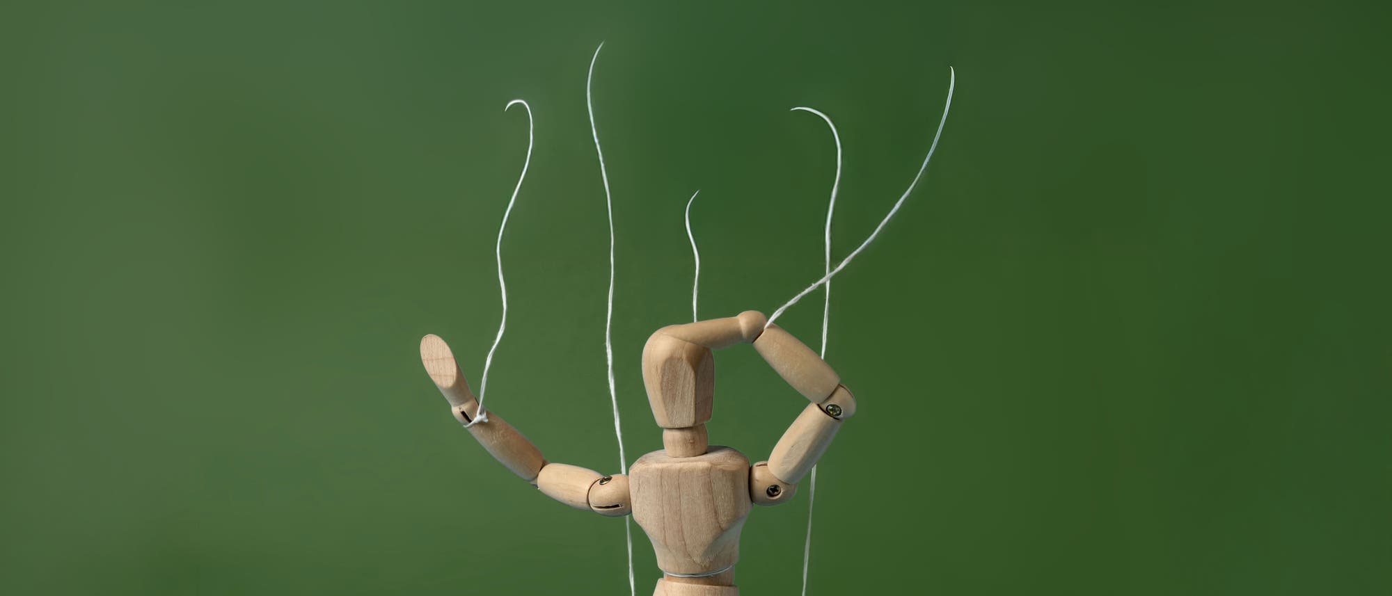 Eine hölzerne Marionette, die an Fäden aufgehängt ist, vor einem hellblauen Hintergrund. Die Marionette hebt einen Arm, während der andere Arm nach unten zeigt. Die Szene vermittelt den Eindruck von Bewegung und Kontrolle.