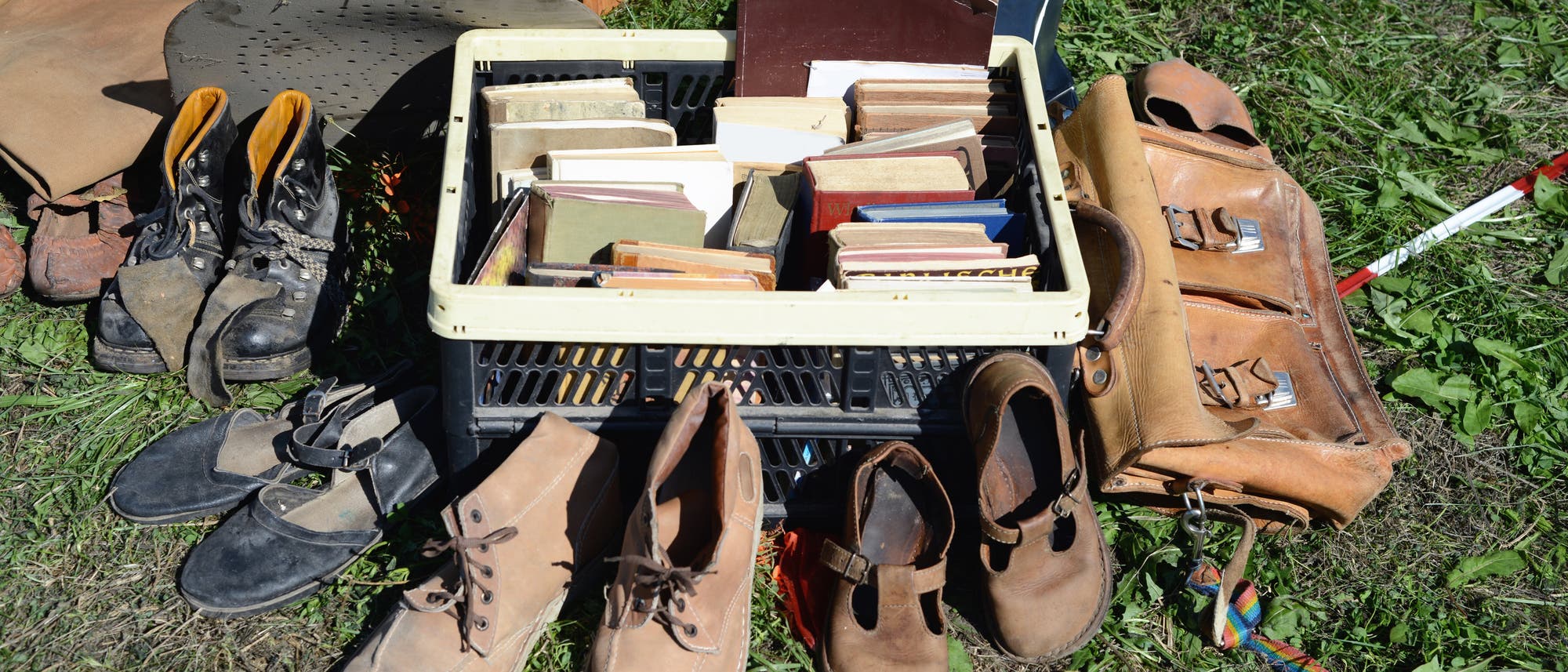 Eine Kiste mit gebrauchten B&uuml;chern steht auf einer Wiese, daneben sind verschiedene Paar Schuhe und eine braune Ledertasche zu sehen. Ein handgeschriebenes Schild auf der Kiste weist darauf hin, dass jedes Buch einen Euro kostet. Im Hintergrund sind weitere Gegenst&auml;nde und ein Teil eines Zeltes sichtbar.