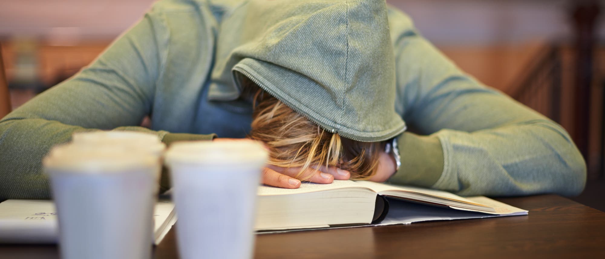 Eine Person in einem grünen Hoodie mit aufgezogener Kapuze sitzt an einem Tisch und hat den Kopf auf ein aufgeschlagenes Buch gelegt, als ob sie schläft oder erschöpft ist. Im Vordergrund stehen zwei Kaffeebecher. Eine Person in einem grünen Hoodie mit aufgezogener Kapuze sitzt an einem Tisch und hat den Kopf auf ein aufgeschlagenes Buch gelegt, als ob sie schläft oder erschöpft ist. Im Vordergrund stehen zwei Kaffeebecher.