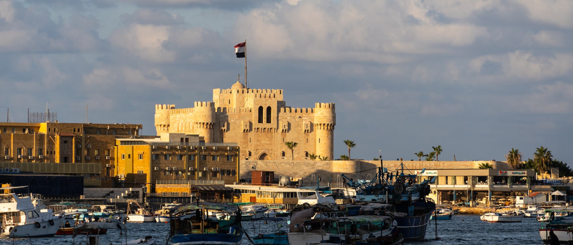 Eine historische Festung mit einer &auml;gyptischen Flagge weht auf einem Turm, umgeben von mehreren Geb&auml;uden. Im Vordergrund befinden sich zahlreiche Boote, die im Wasser vor Anker liegen. Der Himmel ist bew&ouml;lkt, und die Szene wird von warmem Sonnenlicht beleuchtet.