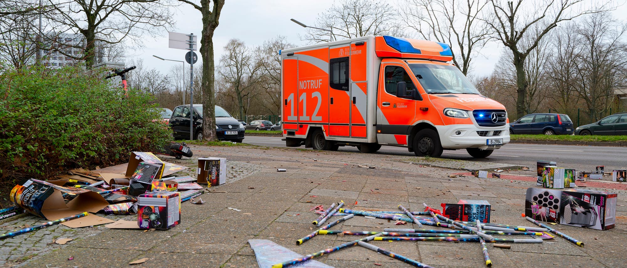 Ein orangefarbener Rettungswagen mit der Aufschrift &raquo;Notruf 112&laquo; steht am Stra&szlig;enrand. Im Vordergrund liegt verstreuter M&uuml;ll, darunter leere Feuerwerksverpackungen und Raketen, auf dem Gehweg. Im Hintergrund sind kahle B&auml;ume und parkende Autos zu sehen. Die Szene wirkt ruhig, ohne sichtbare Personen oder Aktivit&auml;ten.