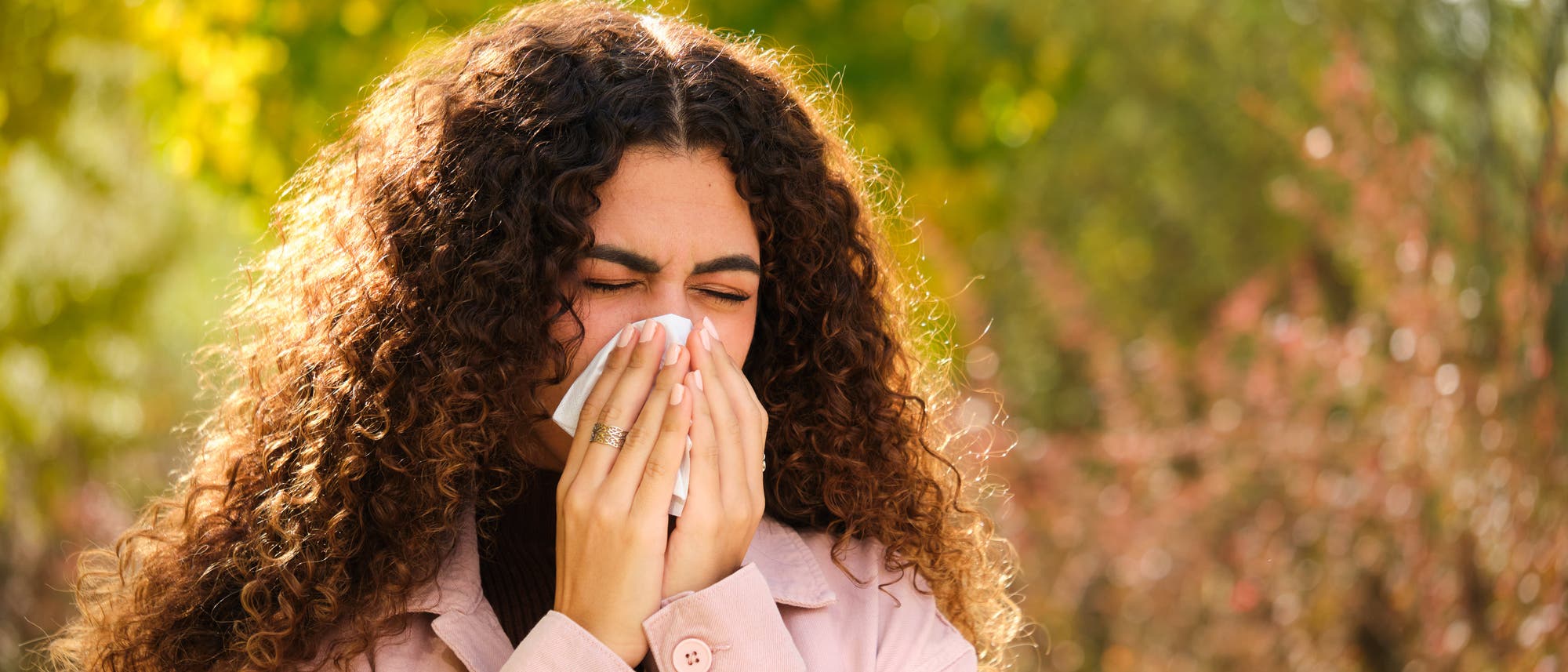 Eine Frau steht im Freien und niest in ein Taschentuch. Sie trägt eine rosa Jacke und hat lockiges, dunkles Haar. Der Hintergrund zeigt unscharfe Bäume und Pflanzen. Eine Frau steht im Freien und niest in ein Taschentuch. Sie trägt eine rosa Jacke und hat lockiges, dunkles Haar. Der Hintergrund zeigt unscharfe Bäume und Pflanzen.