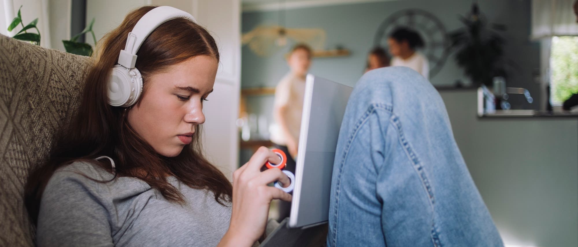 Eine junge Frau liegt halb aufgerichtet auf einem Sofa. Sie trägt weiße Kopfhörer und schaut konzentriert auf ein Tablet, das auf ihrem Bauch steht. In ihren Händen hält sie ein so genanntes Fidget Toy, das der Entspannung dienen soll. Im Hintergrund sind unscharf zwei Personen in einer Küche zu sehen.