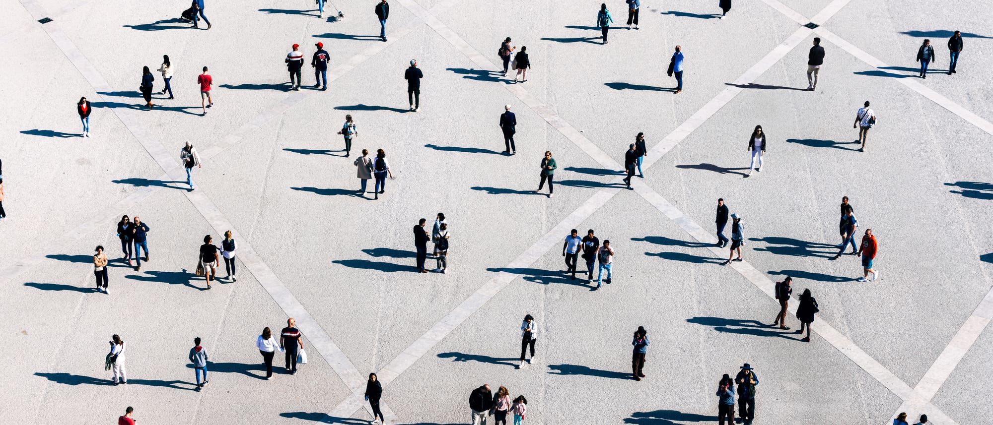 Eine große, offene Fläche mit vielen Menschen, die in kleinen Gruppen oder allein stehen und gehen. Die Szene ist aus der Vogelperspektive aufgenommen, wodurch geometrische Muster auf dem Boden sichtbar werden. Die Personen werfen lange Schatten. Die Kleidung der Menschen ist vielfältig, was auf eine alltägliche, urbane Umgebung schließen lässt. Eine große, offene Fläche mit vielen Menschen, die in kleinen Gruppen oder allein stehen und gehen. Die Szene ist aus der Vogelperspektive aufgenommen, wodurch geometrische Muster auf dem Boden sichtbar werden. Die Personen werfen lange Schatten. Die Kleidung der Menschen ist vielfältig, was auf eine alltägliche, urbane Umgebung schließen lässt.