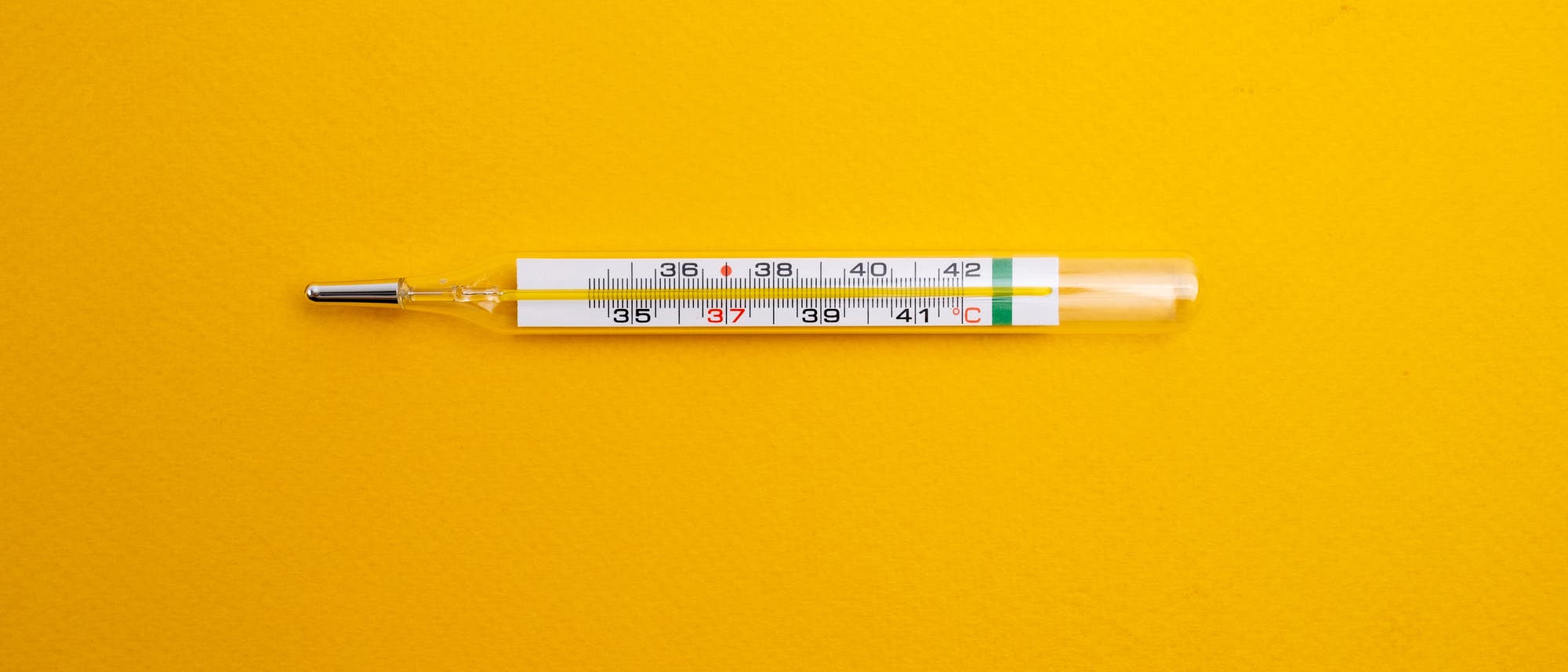Ein Glasthermometer liegt auf einem leuchtend gelben Hintergrund. Die Skala zeigt Temperaturen von 35°C bis 42°C an, wobei der rote Quecksilberfaden bei etwa 37°C steht.