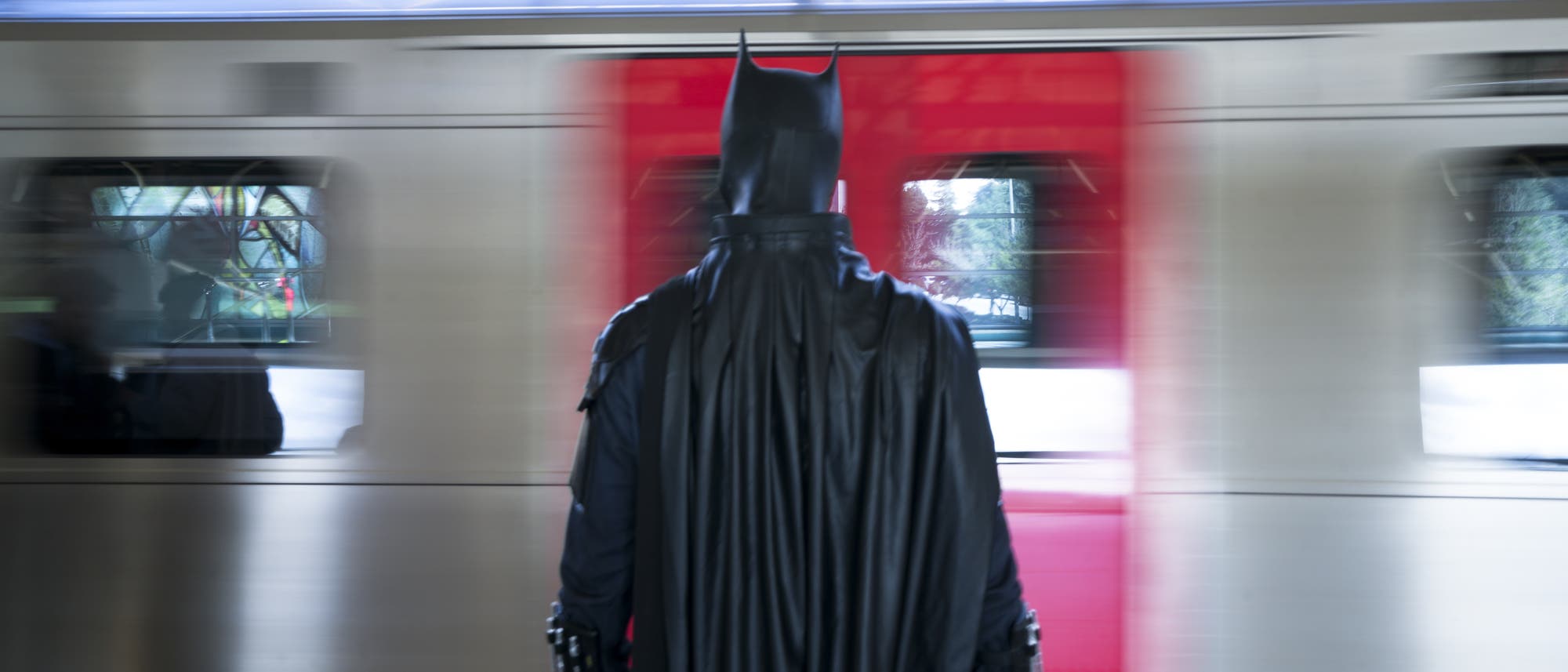 Eine Person in einem Batman-Kostüm steht auf einem Bahnsteig und blickt auf einen vorbeifahrenden Zug. Der Zug ist in Bewegung, was durch die verschwommenen Fenster sichtbar ist. Die Szene spielt sich in einer modernen, überdachten Bahnhofsumgebung ab.