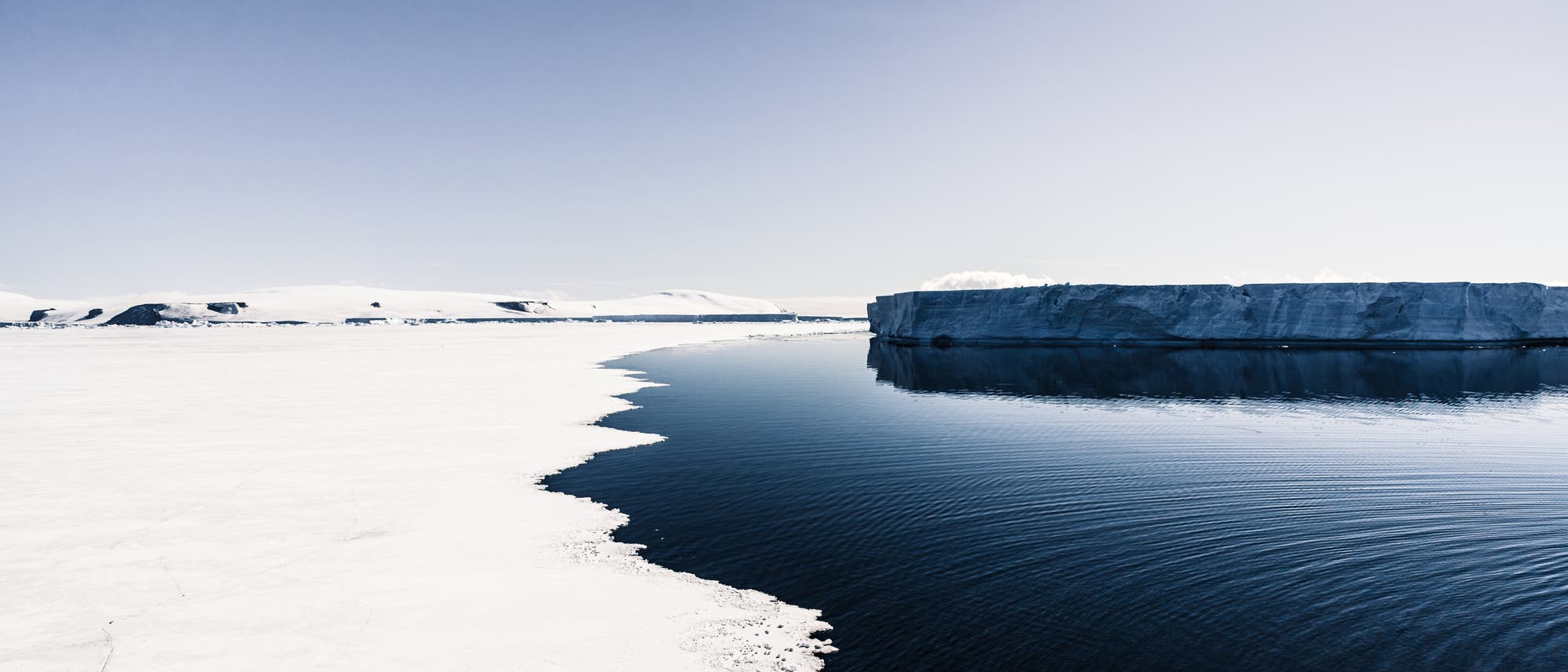 Offenes, dunkles Wasser erstreckt sich zwischen hellem, weißem Eis links im Bild und einem dunklen Eisberg rechts. Der Himmel ist blassblau.