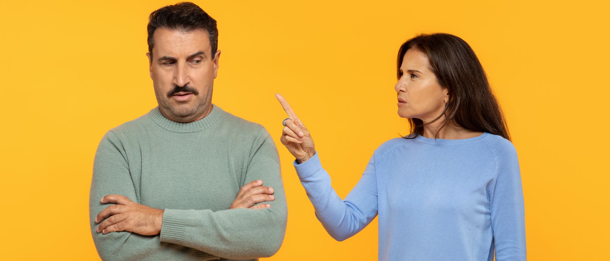 Eine Frau in einem blauen Pullover gestikuliert mit erhobenem Zeigefinger und spricht mit einem Mann in einem gr&uuml;nen Pullover, der die Arme verschr&auml;nkt hat. Der Hintergrund ist einfarbig orange. Die Szene deutet auf eine Diskussion oder Meinungsverschiedenheit hin.
