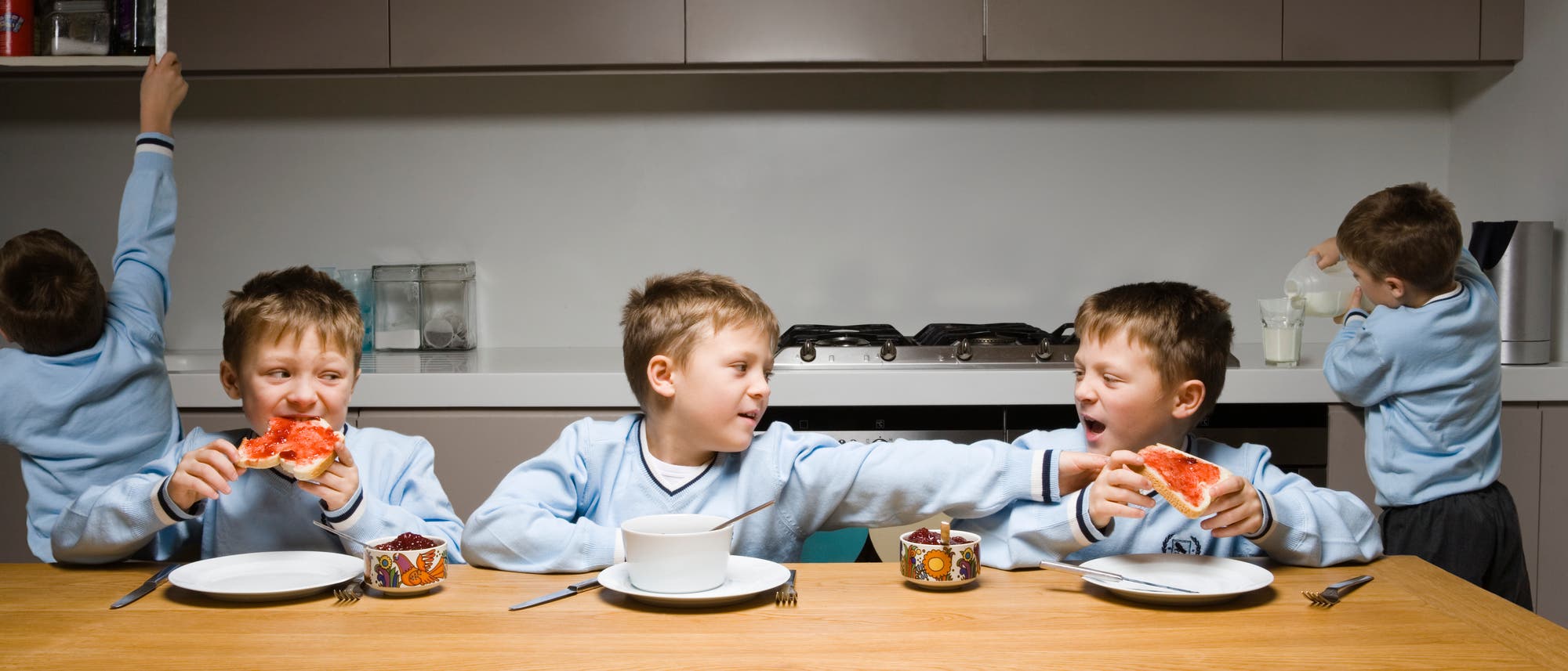 Ein Junge in einem blauen Pullover sitzt an einem Tisch und isst ein Marmeladenbrot. Neben ihm sitzt ein weiterer Junge, der ihm das Brot wegnehmen möchte. Links von ihm isst ein weiterer Junge ebenfalls ein Marmeladenbrot. Auf dem Tisch stehen eine Schüssel, Tassen und Besteck. Im Hintergrund öffnet ein Junge einen Schrank, während ein anderer Milch in ein Glas gießt. Ein Junge in einem blauen Pullover sitzt an einem Tisch und isst ein Marmeladenbrot. Neben ihm sitzt ein weiterer Junge, der ihm das Brot wegnehmen möchte. Links von ihm isst ein weiterer Junge ebenfalls ein Marmeladenbrot. Auf dem Tisch stehen eine Schüssel, Tassen und Besteck. Im Hintergrund öffnet ein Junge einen Schrank, während ein anderer Milch in ein Glas gießt.