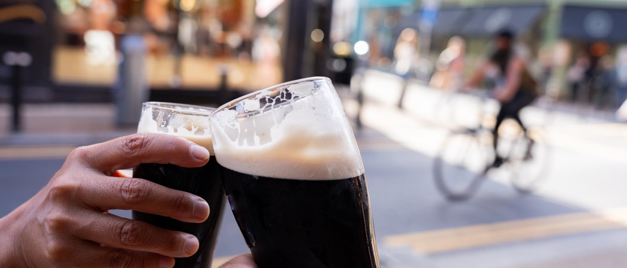 Zwei Personen stoßen im Freien bei gutem Wetter mit einem Guinness-Bier an, im Hintergrund sind Geschäfte und Passanten in Unschärfe zu sehen.