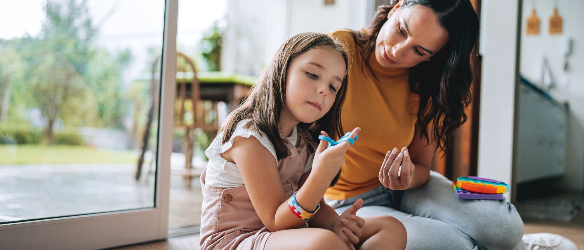 Eine Frau und ein Mädchen sitzen auf dem Boden in einem hellen Raum mit großen Fenstern. Das Mädchen hält ein Spielzeug in der Hand, sie betrachtet es intensiv und wirkt in sich gekehrt. Die Frau schaut aufmerksam zu und beugt sich zu ihr. Beide tragen lässige Kleidung. Im Hintergrund ist ein Tisch mit Stühlen und ein grüner Garten zu sehen. Die Szene vermittelt eine ruhige und konzentrierte Atmosphäre.