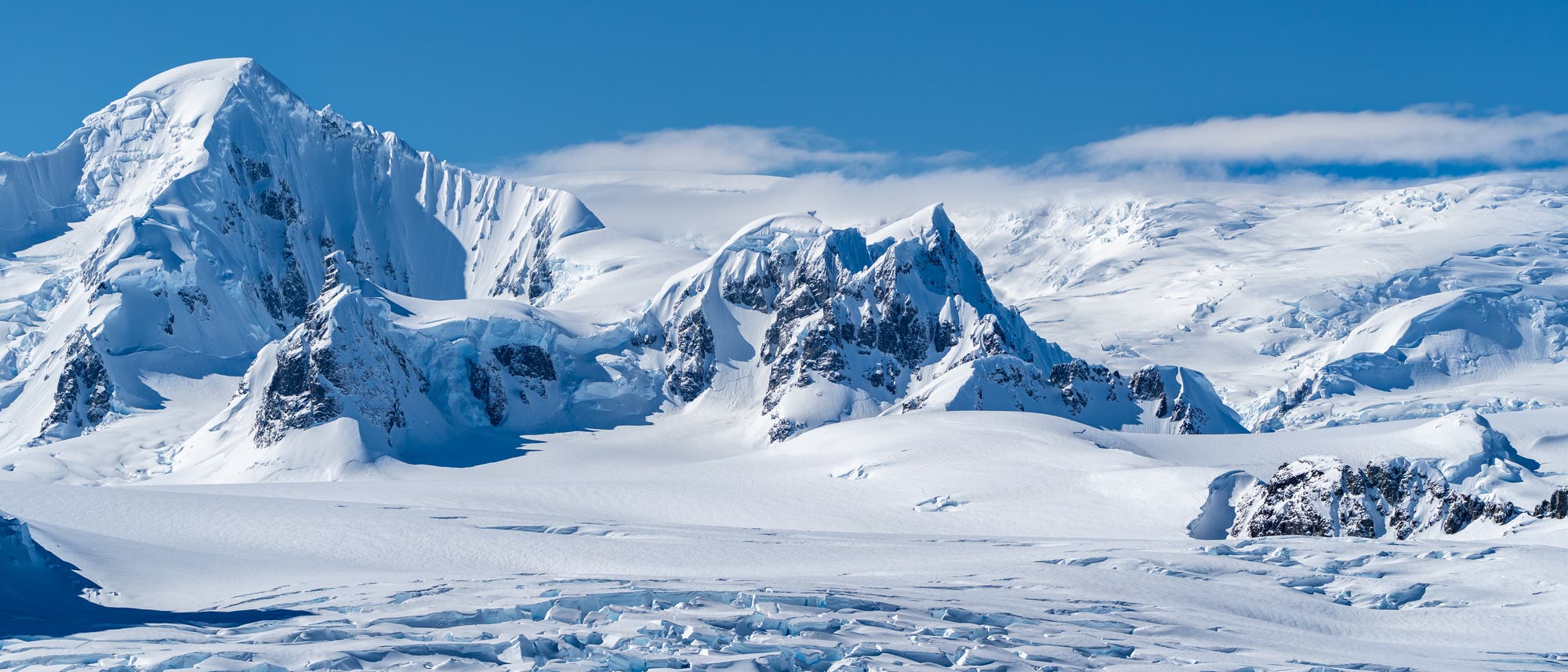 Verschneite Berglandschaft mit schroffen Gipfeln und Gletschern unter einem klaren blauen Himmel. Die Szene zeigt eine beeindruckende Eislandschaft mit scharfen Felsformationen und weiten Schneefeldern. Im Vordergrund sind zerkl&uuml;ftete Gletscherstrukturen sichtbar.