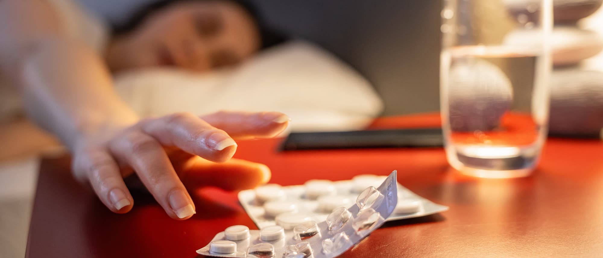 Eine Person liegt im Bett und streckt die Hand nach einem Blister mit Tabletten auf einem Nachttisch aus. Neben den Tabletten steht ein Glas Wasser. Die Szene ist in einem Schlafzimmer bei gedämpftem Licht.
