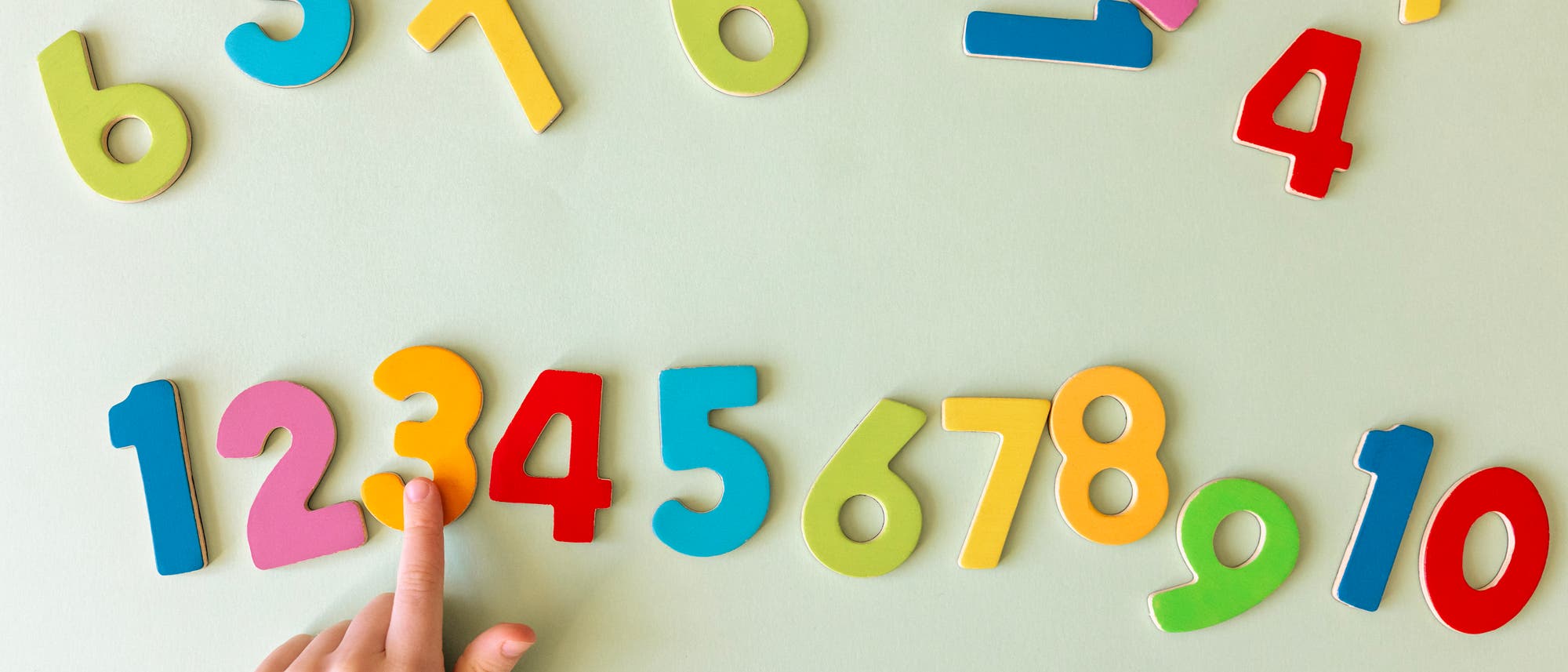 Eine Kinderhand zeigt auf bunte, magnetische Zahlen auf einer hellgrünen Oberfläche. Die Zahlen sind in verschiedenen Farben wie Rot, Blau, Gelb und Grün dargestellt und reichen von 1 bis 10. Die Anordnung der Zahlen ist spielerisch und unregelmäßig. Eine Kinderhand zeigt auf bunte, magnetische Zahlen auf einer hellgrünen Oberfläche. Die Zahlen sind in verschiedenen Farben wie Rot, Blau, Gelb und Grün dargestellt und reichen von 1 bis 10. Die Anordnung der Zahlen ist spielerisch und unregelmäßig.
