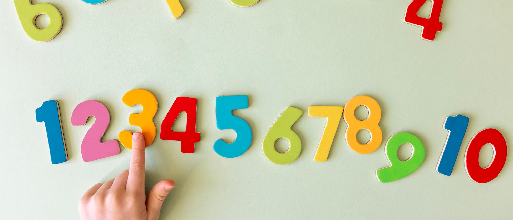 Eine Kinderhand zeigt auf eine Reihe bunter Zahlen von 1 bis 10, die auf einem hellgrünen Hintergrund angeordnet sind. Die Zahlen sind aus Holz und in verschiedenen Farben wie Blau, Rot, Gelb und Grün gestaltet. Oben im Bild sind weitere Zahlen verstreut.