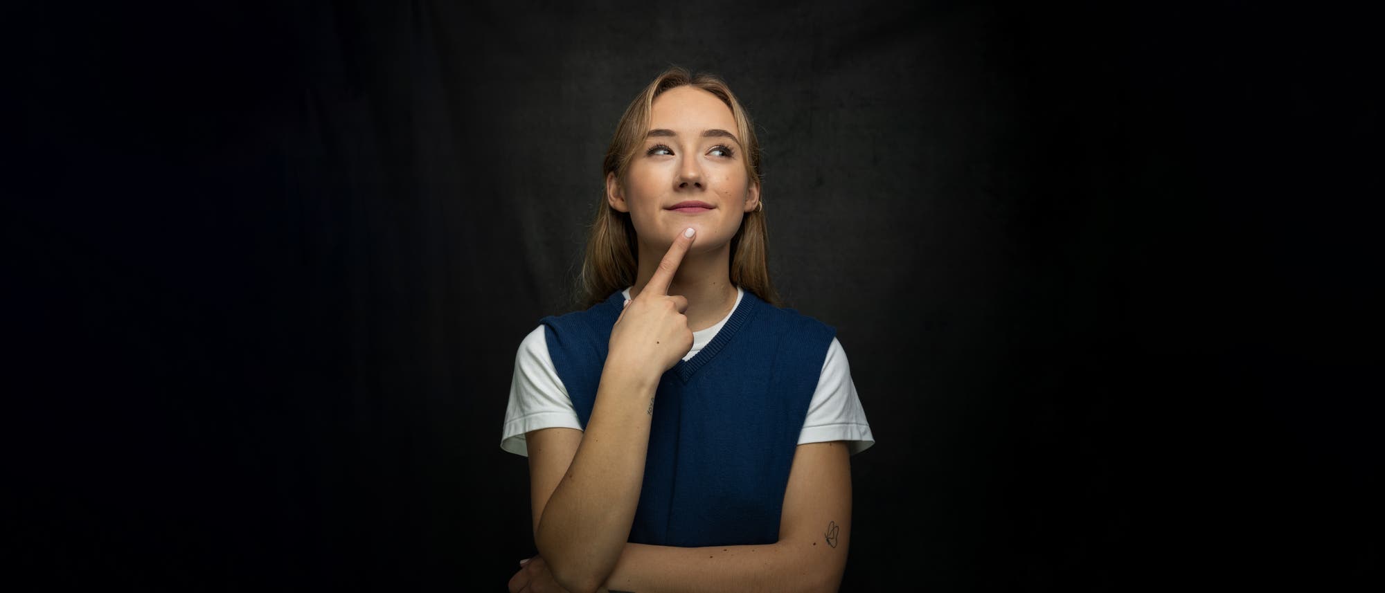 Eine Frau steht vor einem dunklen Hintergrund und schaut nachdenklich nach oben, während sie mit einem Finger an ihr Kinn tippt. Sie trägt ein blaues Oberteil über einem weißen T-Shirt. Die Szene vermittelt einen Moment des Nachdenkens oder der Überlegung.