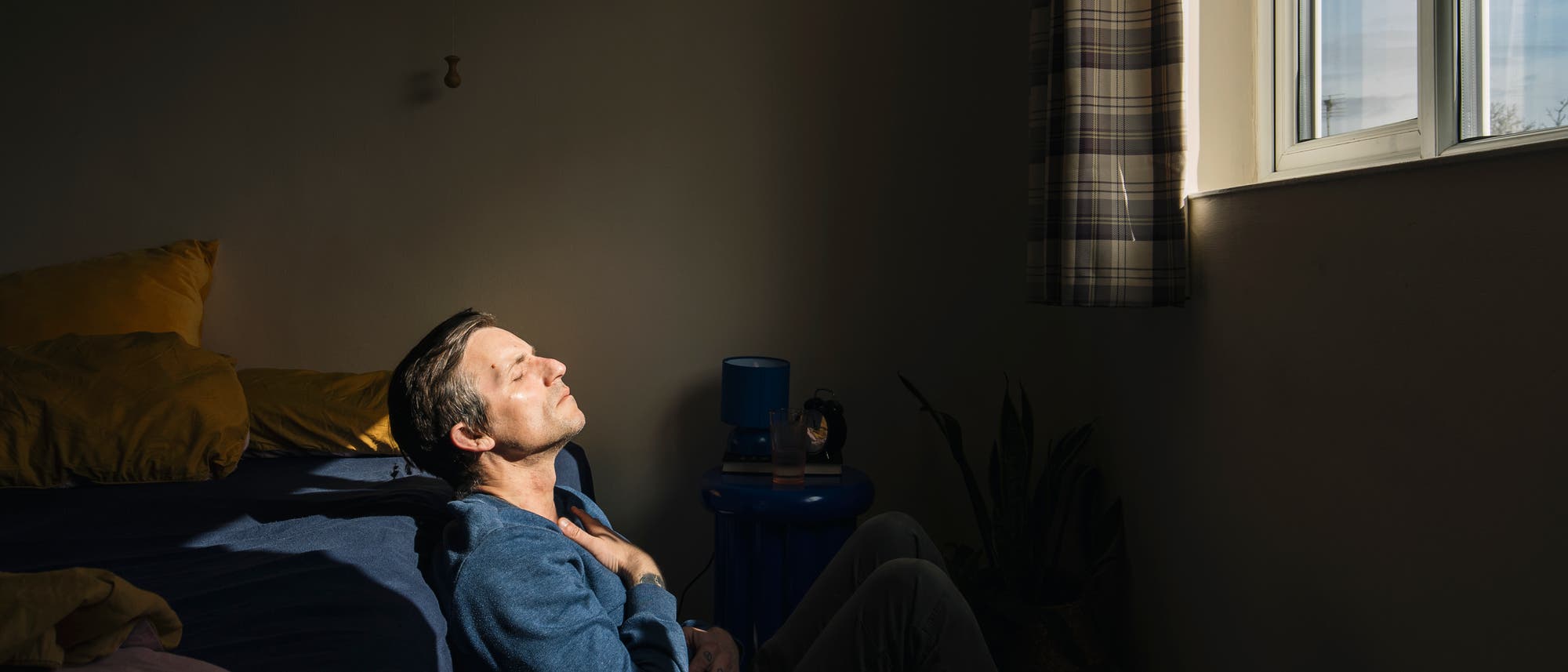 Eine Person sitzt auf dem Boden eines Schlafzimmers, lehnt sich an das Bett und genie&szlig;t das Sonnenlicht, das durch ein Fenster f&auml;llt. Die Person tr&auml;gt einen blauen Pullover und hat die Augen geschlossen, w&auml;hrend sie eine Hand auf die Brust legt. Neben dem Bett steht ein kleiner Tisch mit einer Lampe und B&uuml;chern. Die Atmosph&auml;re wirkt ruhig und entspannt.