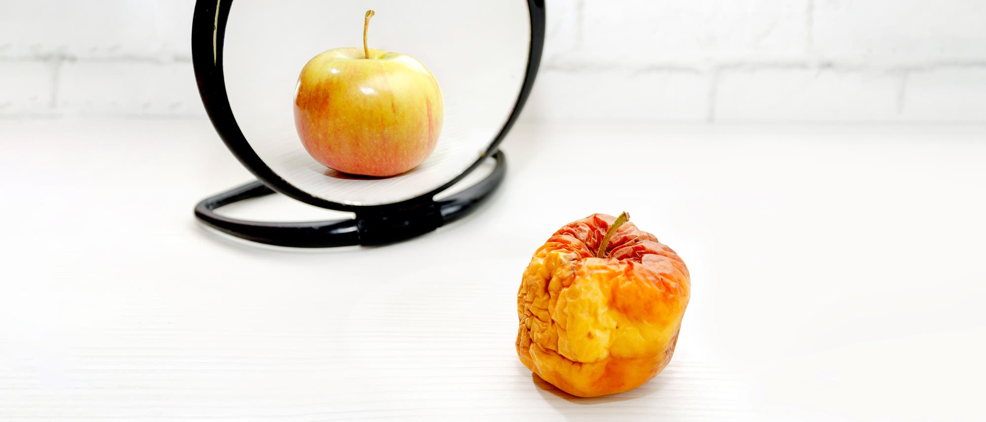 Ein schrumpeliger, alter Apfel steht vor einem Spiegel auf einem weißen Tisch. Im Spiegelbild erscheint der Apfel frisch und makellos. Ein schrumpeliger, alter Apfel steht vor einem Spiegel auf einem weißen Tisch. Im Spiegelbild erscheint der Apfel frisch und makellos.