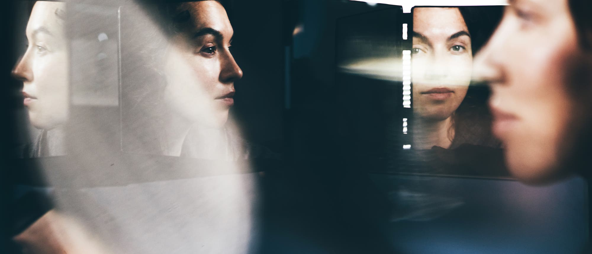 Eine Frau wird in mehreren Spiegeln reflektiert, wodurch verschiedene Ansichten ihres Gesichts sichtbar werden. Die Szene ist k&uuml;nstlerisch beleuchtet, mit einem Fokus auf den Ausdruck und die Reflexionen. Die Spiegel erzeugen eine interessante visuelle Komposition, die Tiefe und Perspektive betont.